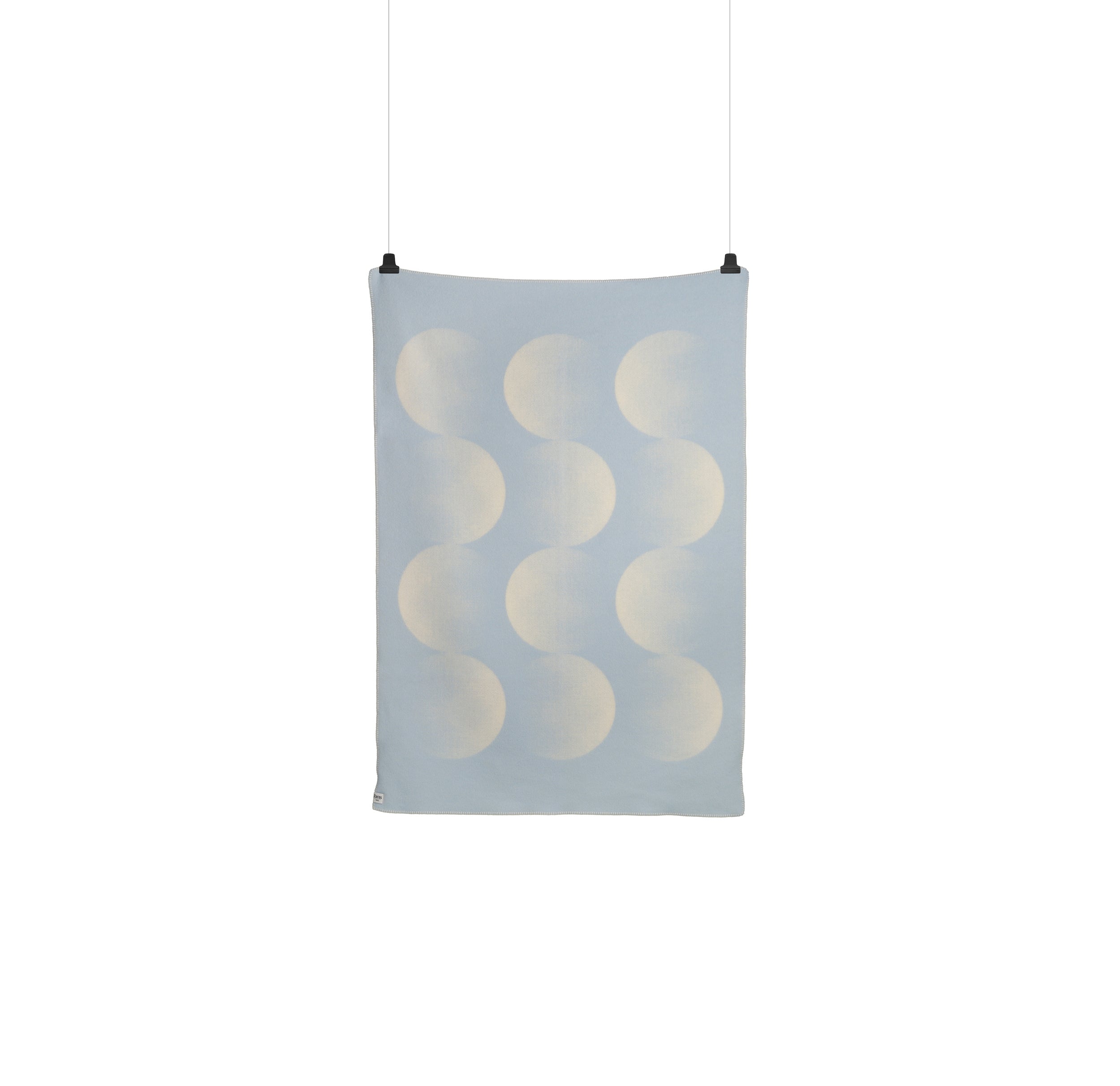 The Moon Large Throw by Roros, made from Norwegian lambswool, is draped against a white backdrop. This light blue rectangular textile showcases crescent-like overlapping patterns in lighter shades, resembling celestial hues and is held up with clothespins.