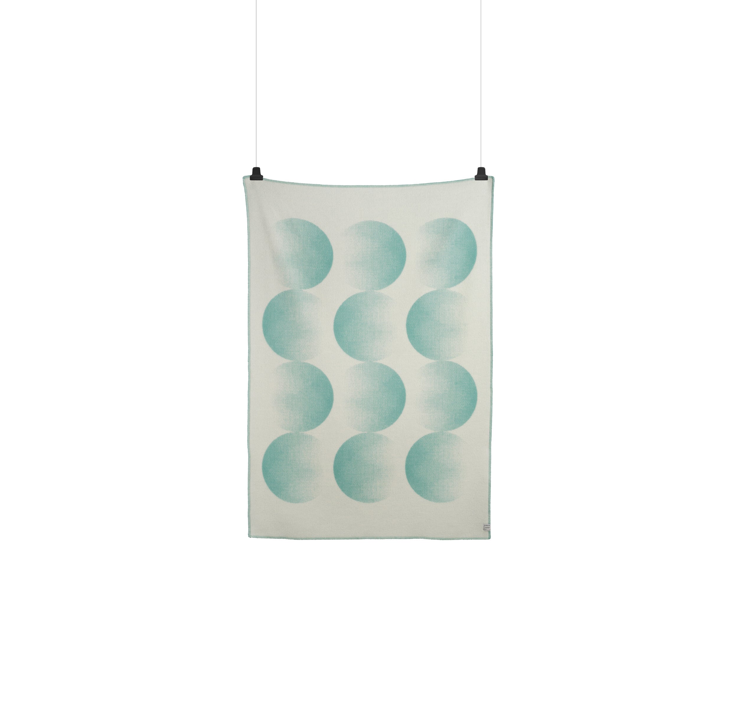 The Roros Moon Large Throw by Clara von Zweigbergk features greenish-blue crescent shapes on beige Norwegian lambswool. It hangs from two black clips in three rows and four columns, creating a wavy design against a white background.
