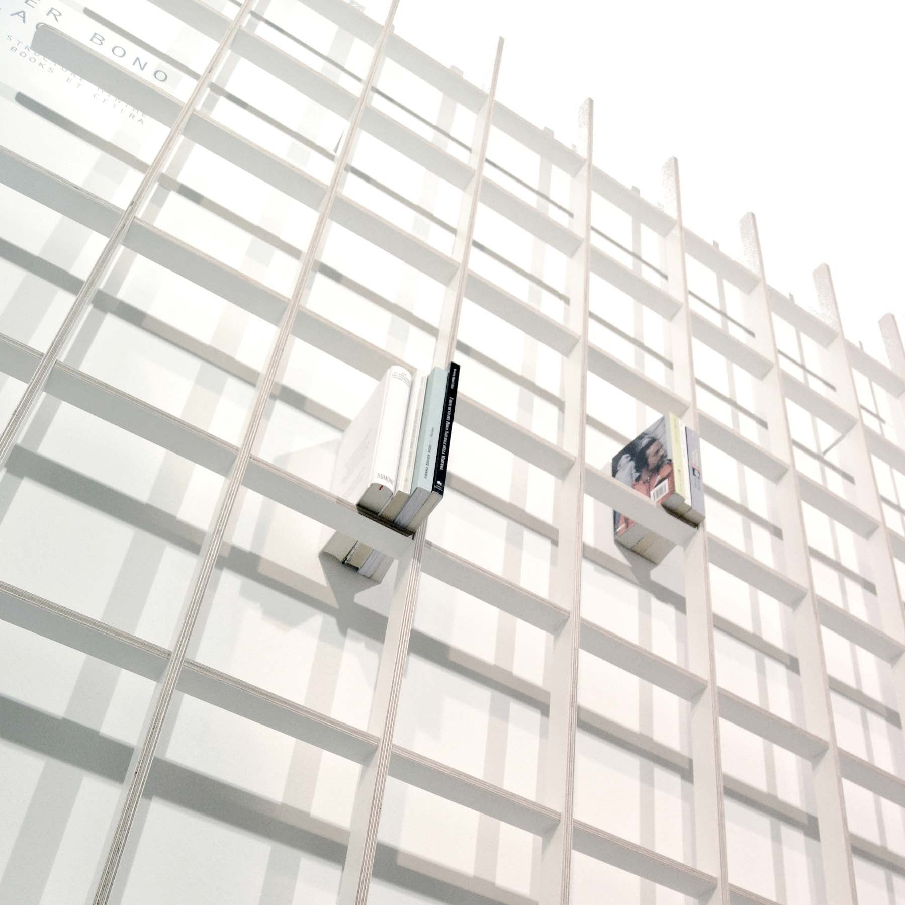 The SCHLAGSEITE bookshelf by Martin Breuer Bono showcases a minimalist, sculptural grid design. It holds a few scattered books, emphasizing its geometric style and clean lines. The angle creates an illusion of the shelves receding into the distance.