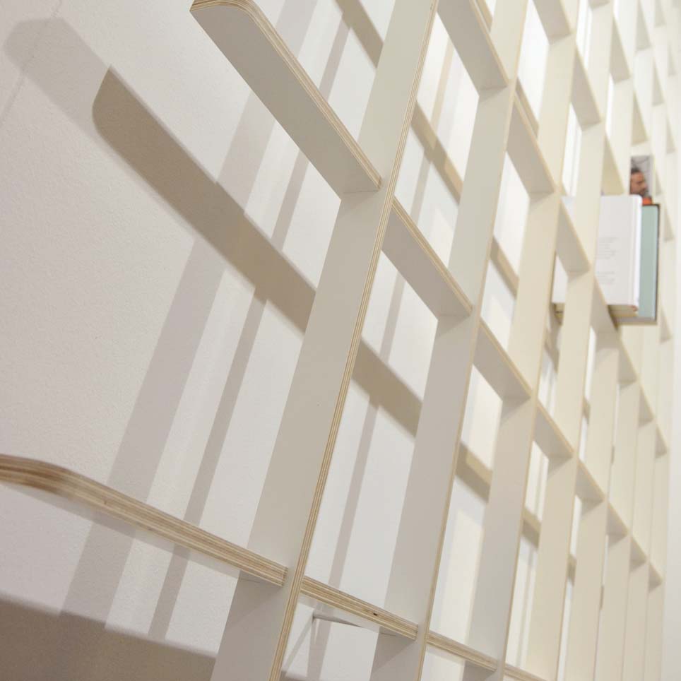 The SCHLAGSEITE by Martin Breuer Bono is a modern, white, grid-like structure with square openings. Its sculptural design casts shadows on a plain wall, while one slot reveals a hint of minimalist functionality with a book section visible, resembling an artistic bookshelf.