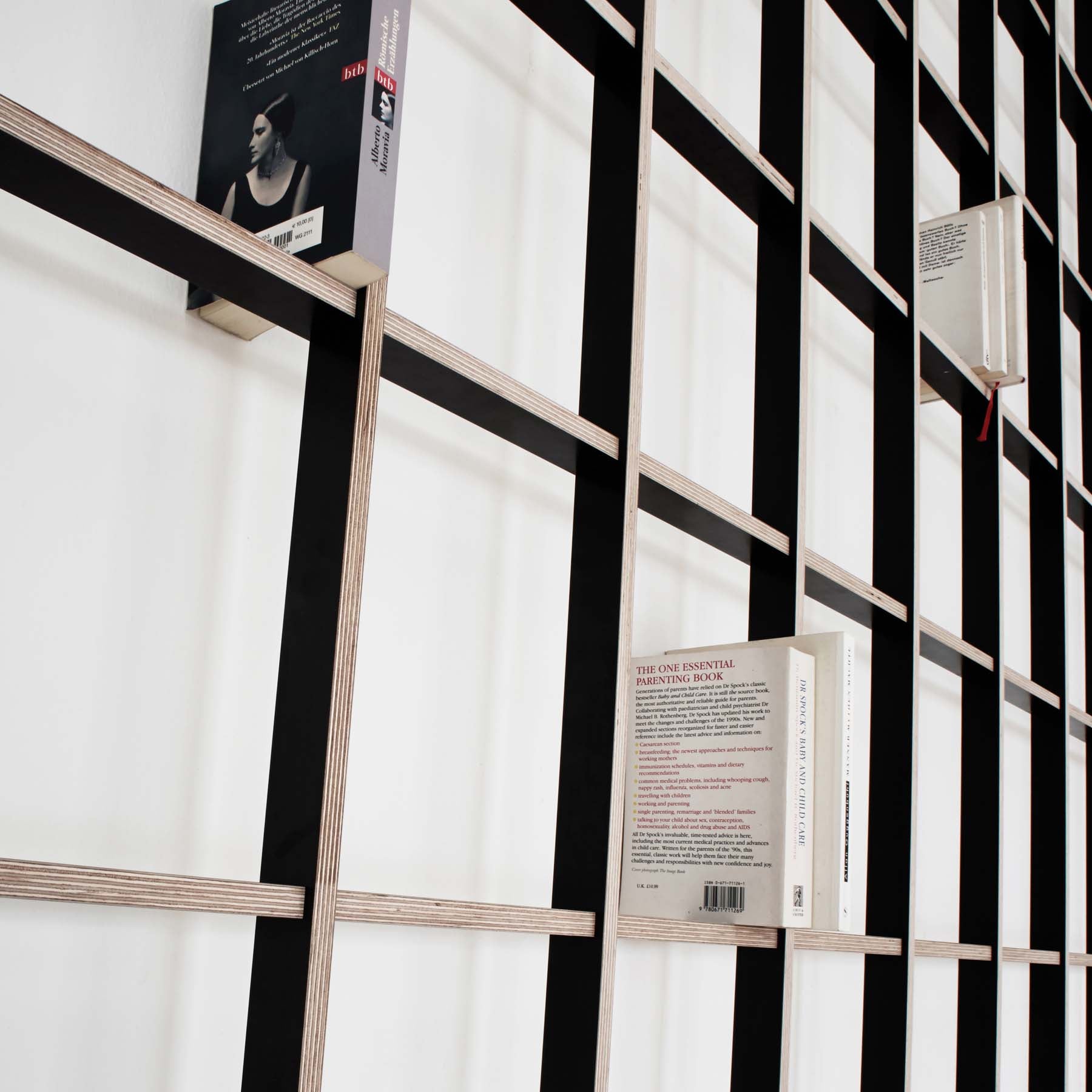 The SCHLAGSEITE bookshelf by Martin Breuer Bono features a modern, minimalist geometric black grid design. Mounted on a white wall, it showcases its sculptural form with books arranged both horizontally and vertically, revealing either covers or spines.
