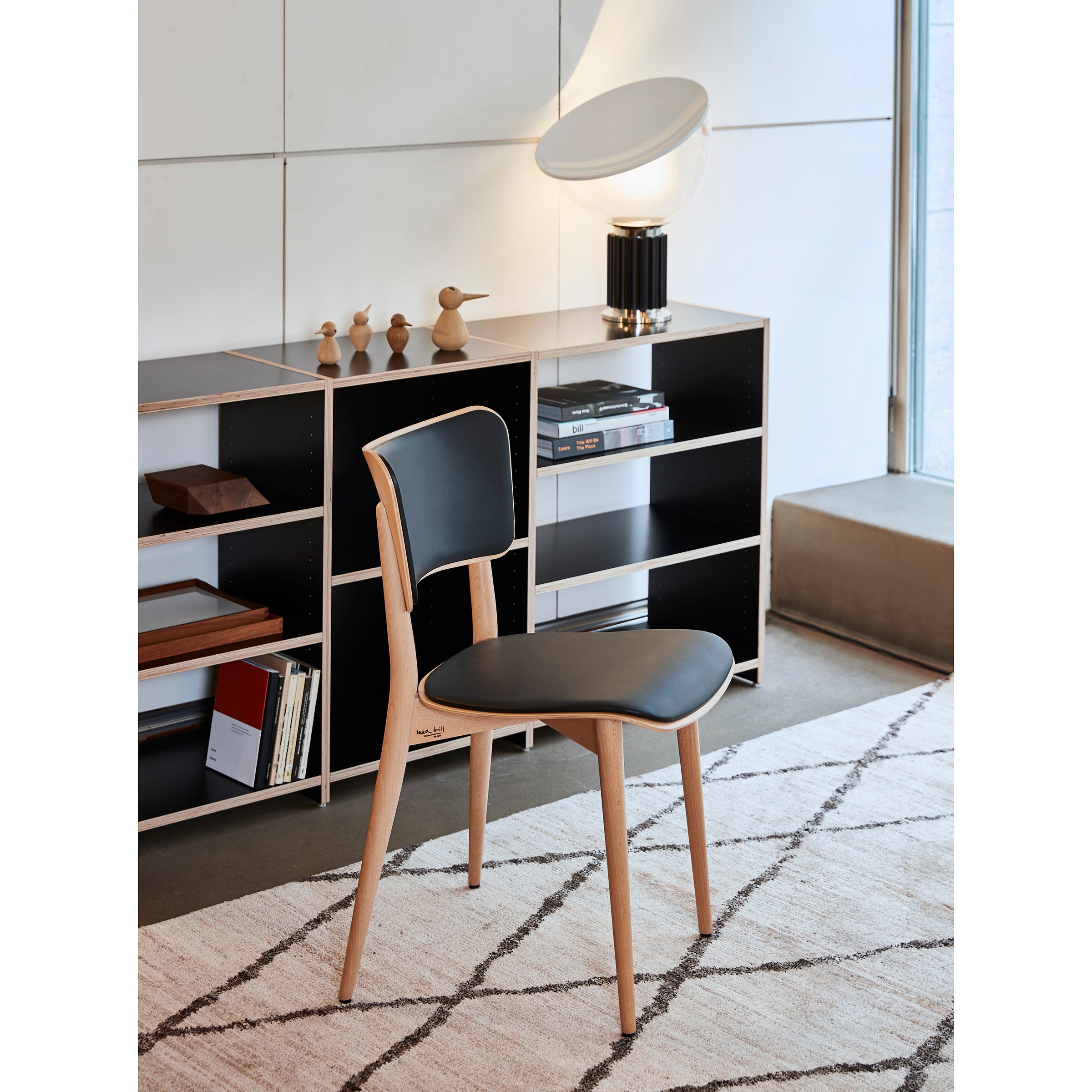 A modern room showcases a WB Form Kreuzzargenstuhl Upholstered chair with black seat and backrest. An open geometric bookshelf behind holds decorative items, books, and a unique lamp. A patterned rug adds floor texture.