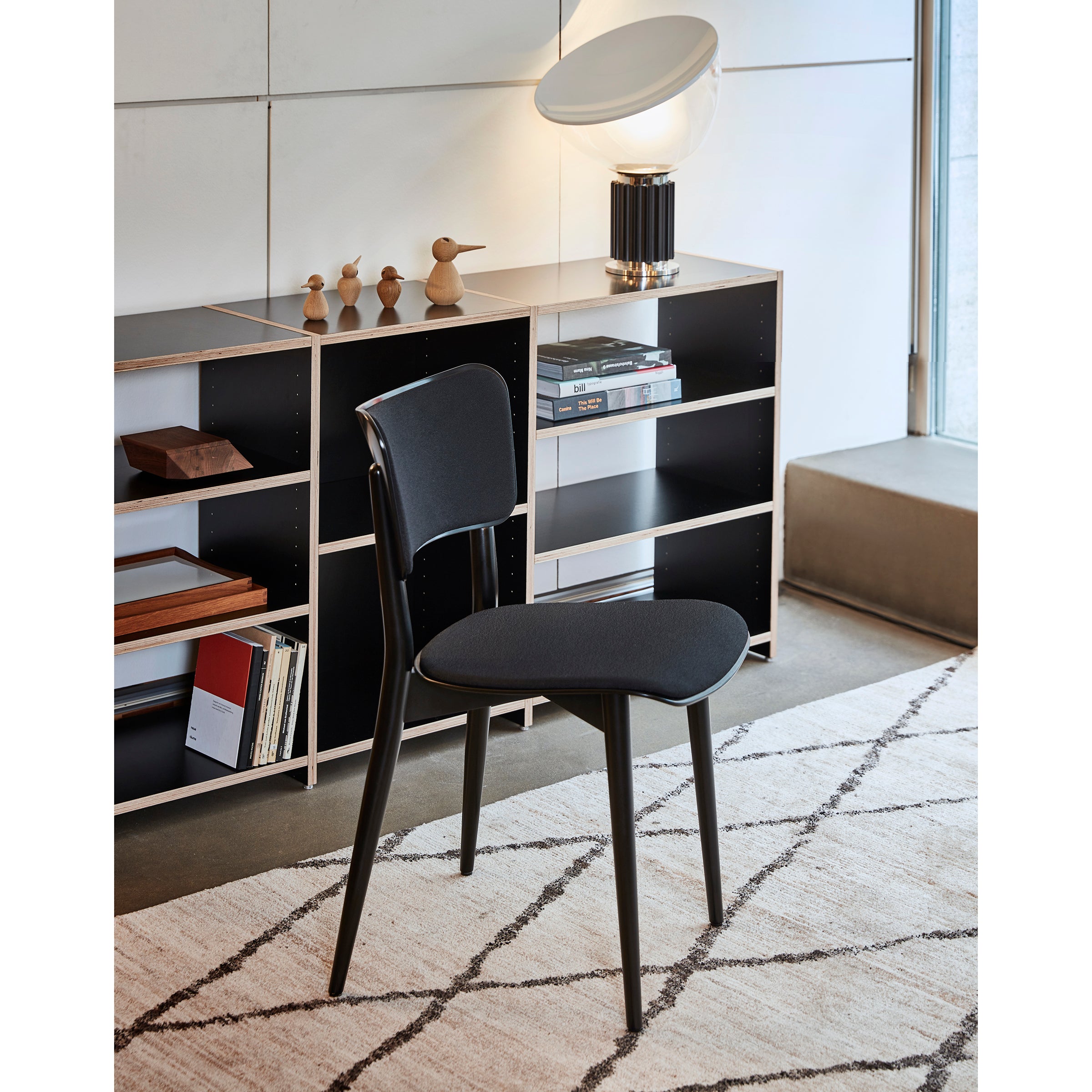 A modern black WB Form Kreuzzargenstuhl Upholstered chair rests on a patterned rug in a stylish room. Behind it are black shelves holding books, decorative wooden birds, and a round-shaded lamp. A large window to the right floods in natural light, accentuating diverse upholstery options for added flair.