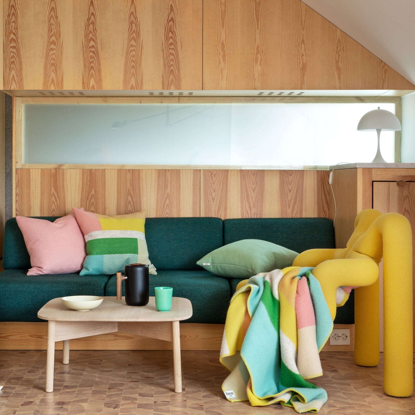 A cozy living room features a green sofa with pastel pillows and a modern, curvy yellow chair draped in the Mikkel Large Throw by Roros. A wooden coffee table with a black pitcher and green cup sits in front, surrounded by warm wood paneling.