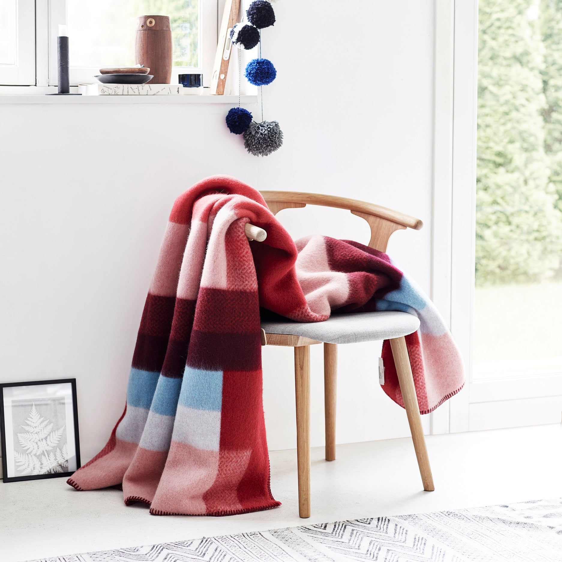 In a bright room, a Mikkel Large Throw by Roros, made of Norwegian wool with colorful stripes, is draped over a wooden chair with a light cushion. A window with potted plants and decorative pom-poms, alongside a framed picture and book on the floor, evokes Bauhaus-inspired design by Kristine Five Melvær.