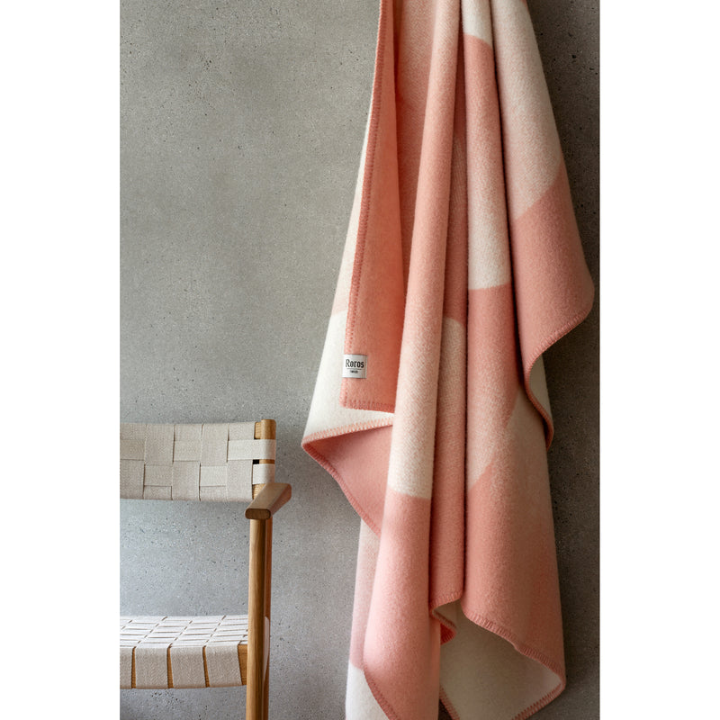 A woven wood chair with a white seat is next to a soft, pink and white checkered Moon Large Throw by Roros, made from luxurious Norwegian lambswool, hanging on a gray textured wall.