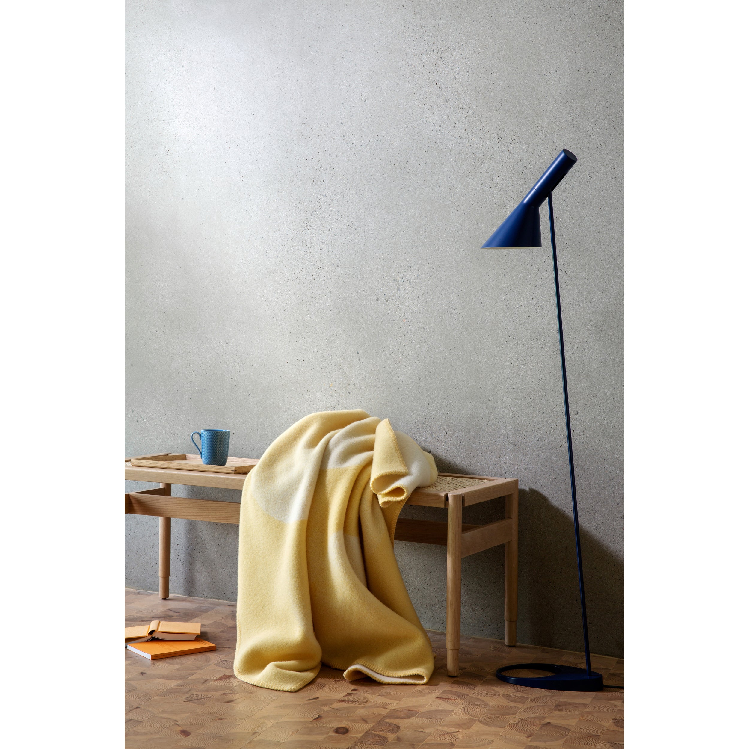 A wooden bench adorned with a Roros Moon Large Throw is set beside a modern floor lamp. A blue mug rests on the bench, while an open notebook is on the floor. The scenes minimalist charm is enhanced by a plain, light gray textured wall in the background.