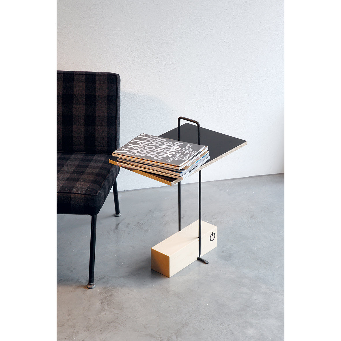 The Abgemahnt side table by Moormann, with a black tabletop and thin metal legs on a rectangular wooden base, complements the black plaid chair. A stack of magazines rests on it, evoking a Nils Holger Moormann aesthetic on a light gray floor.