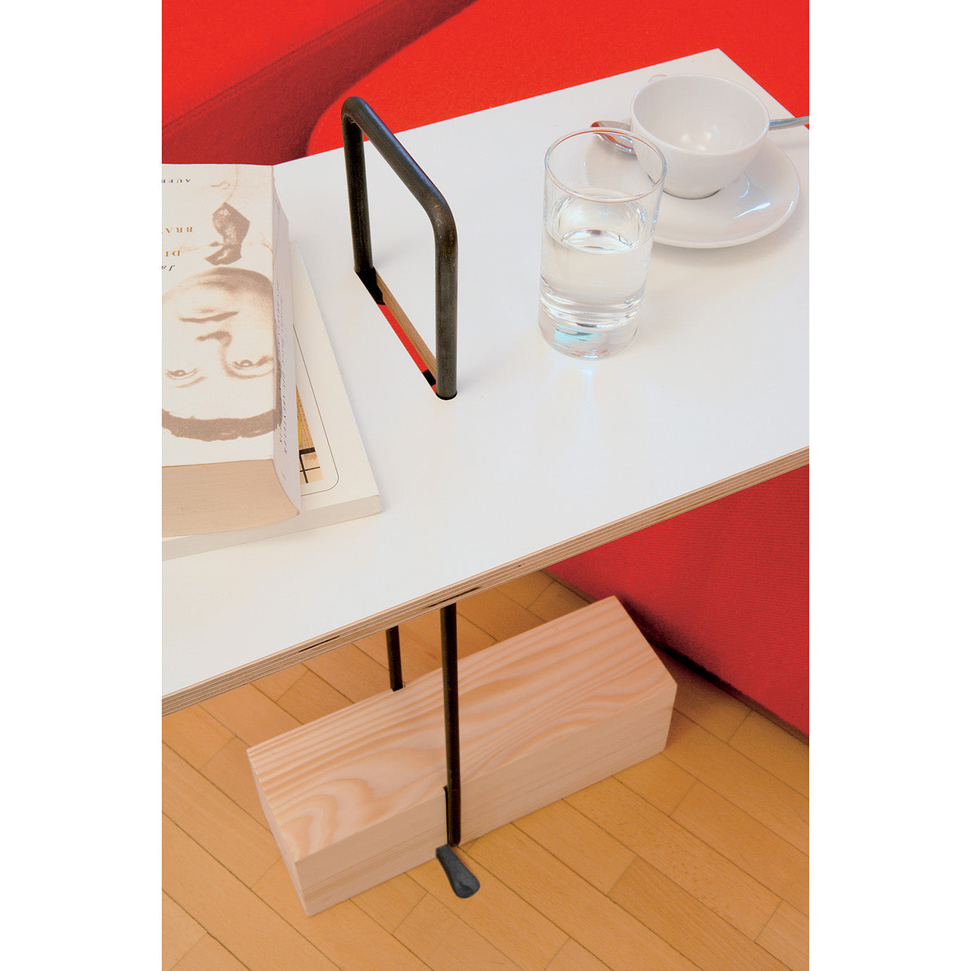 The Abgemahnt side table by Moormann features a striking design with a white tabletop and wooden base. It holds an upside-down book showing a black and white portrait, alongside a glass of water, and a white cup and saucer. This elegant piece sits next to a red couch on the wooden floor.