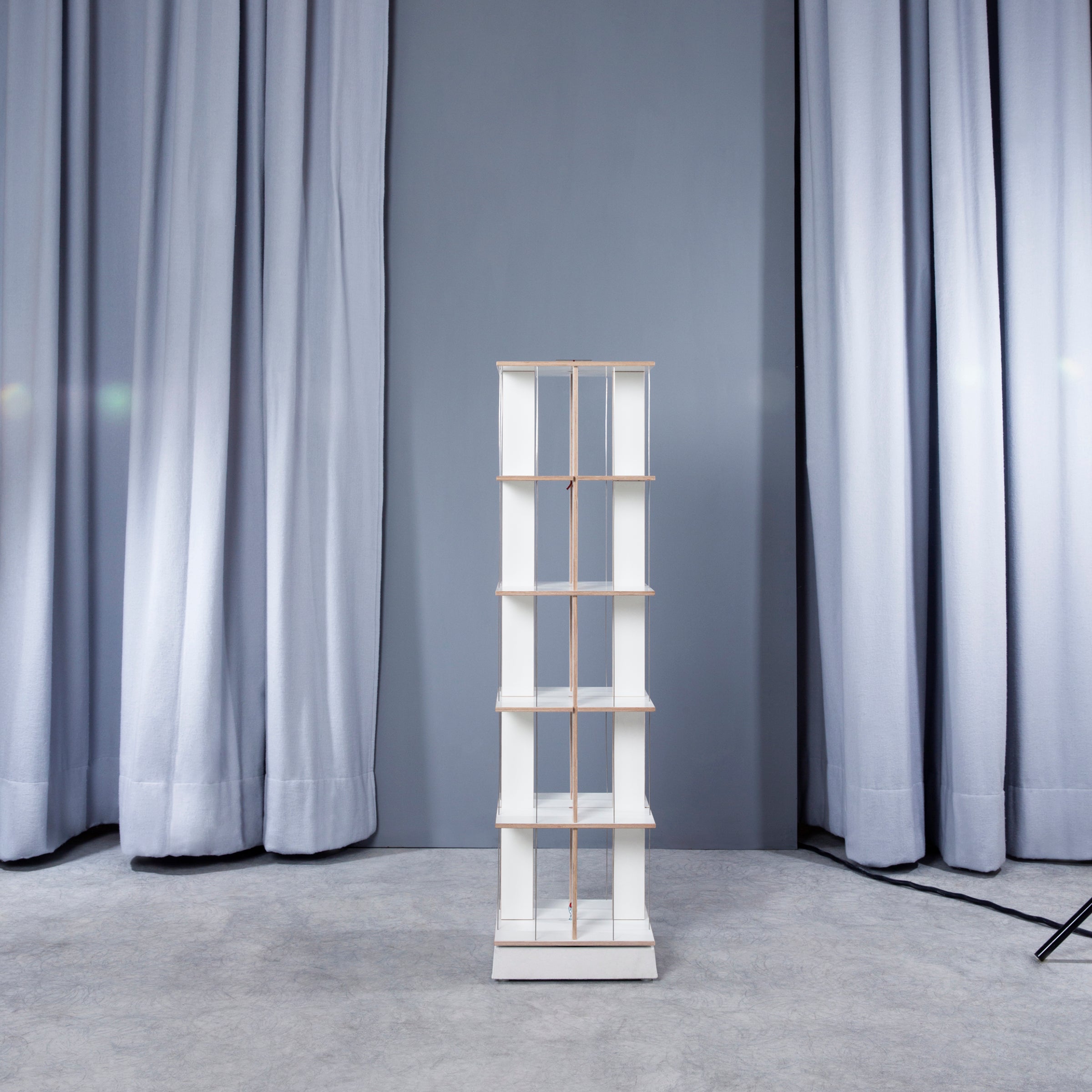 A Moormann Buchstabler rack, known for its space-saving design, features four white shelves supported by thin wooden rods. Its set against long light blue curtains, with a gray floor that enhances the minimalist elegance.