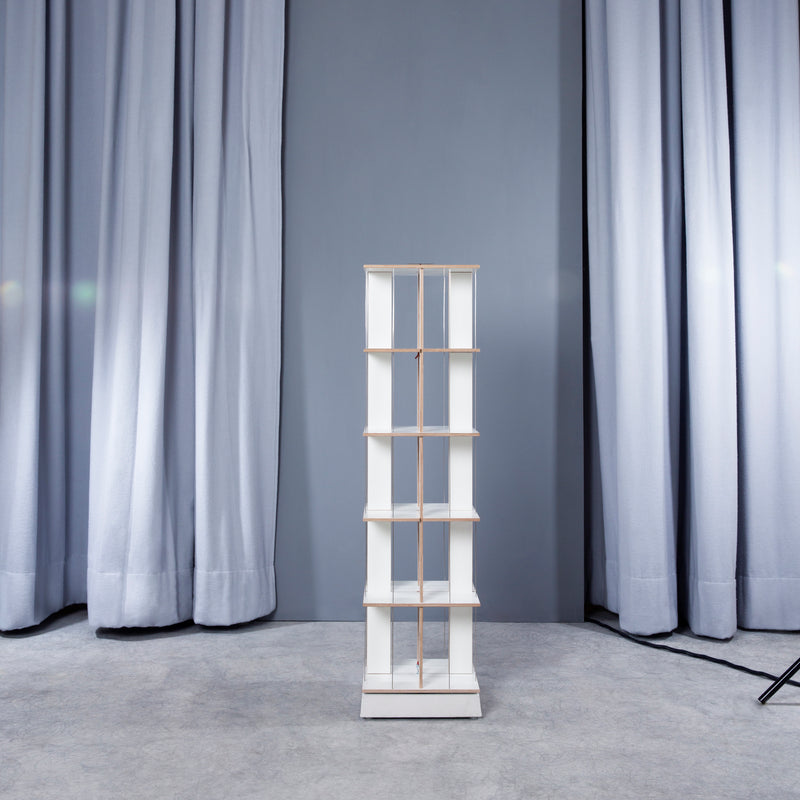 A Moormann Buchstabler rack, known for its space-saving design, features four white shelves supported by thin wooden rods. Its set against long light blue curtains, with a gray floor that enhances the minimalist elegance.