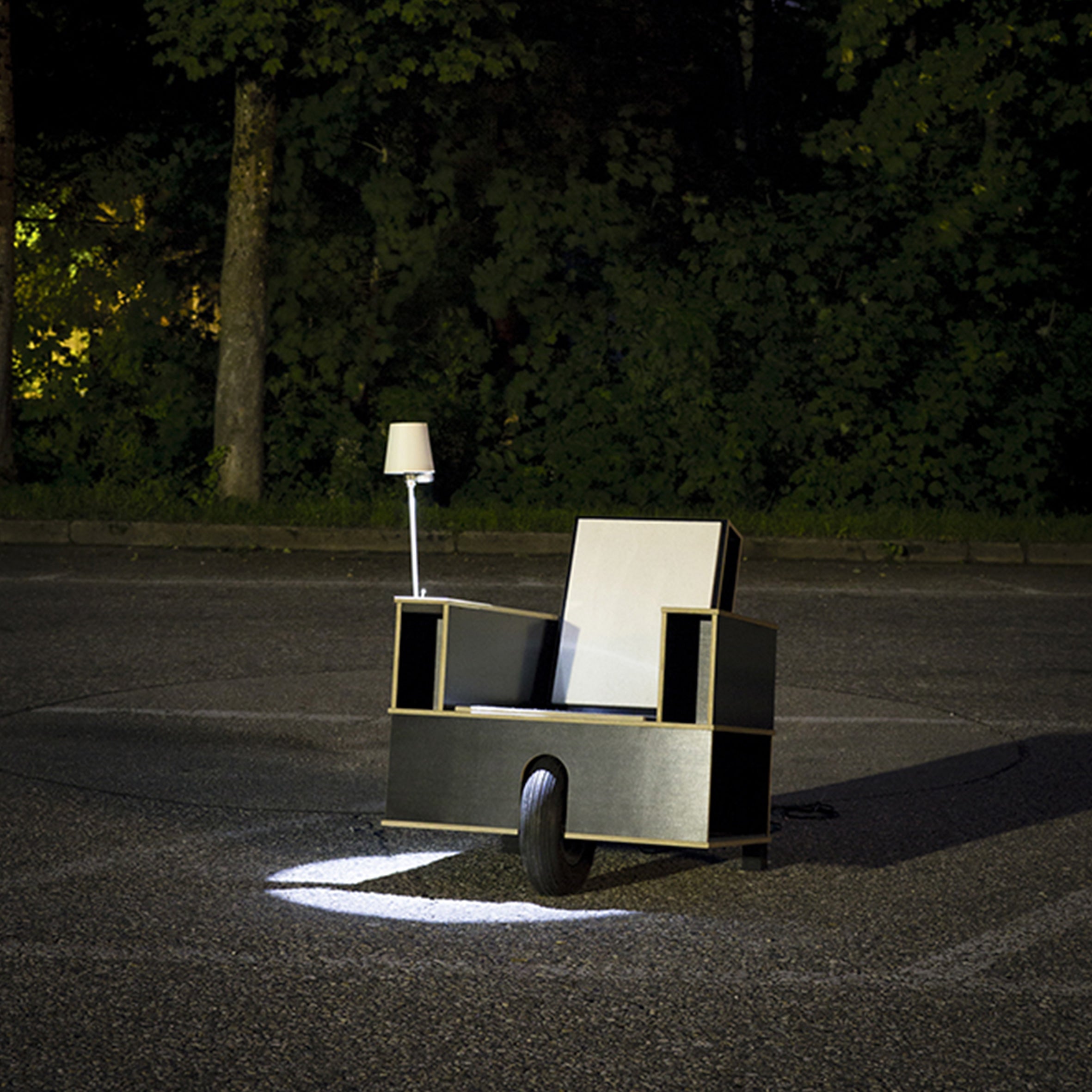The Moormann Bookinist is a modern, boxy reading chair with a built-in lamp. It features a single wheel and stands in a dimly lit outdoor area surrounded by trees, casting a spotlight for an intriguing and minimalist design.