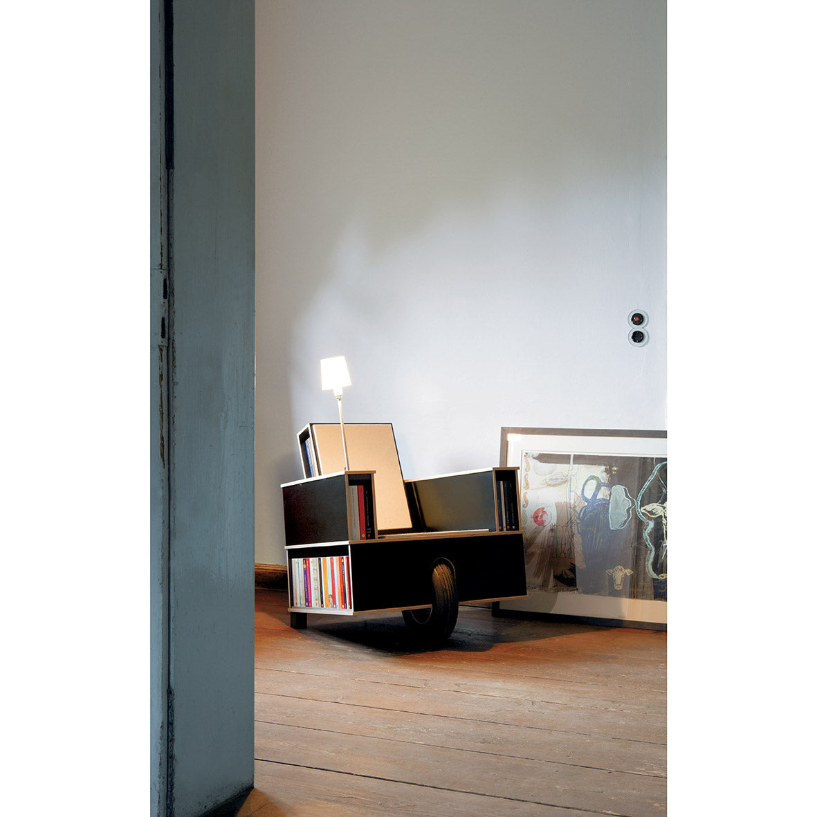 The Moormann Bookinist is a modern reading chair with built-in book storage, featuring shelves filled with books and a small lamp on one armrest. Placed against a white wall on the wooden floor, it complements a framed abstract art piece for the perfect literary nook.