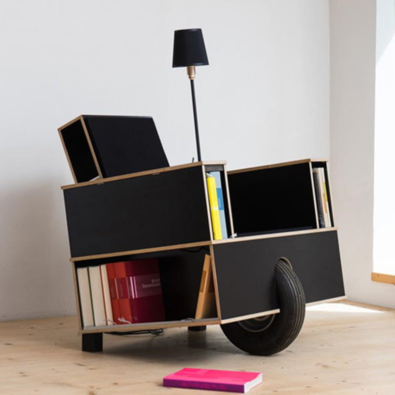 The Moormann Bookinist is a black, wheelbarrow-inspired chair with side shelves for book storage and a small attached lamp. It features a front wheel and sits on a wooden floor, with a pink book nearby to create an inviting reading corner.
