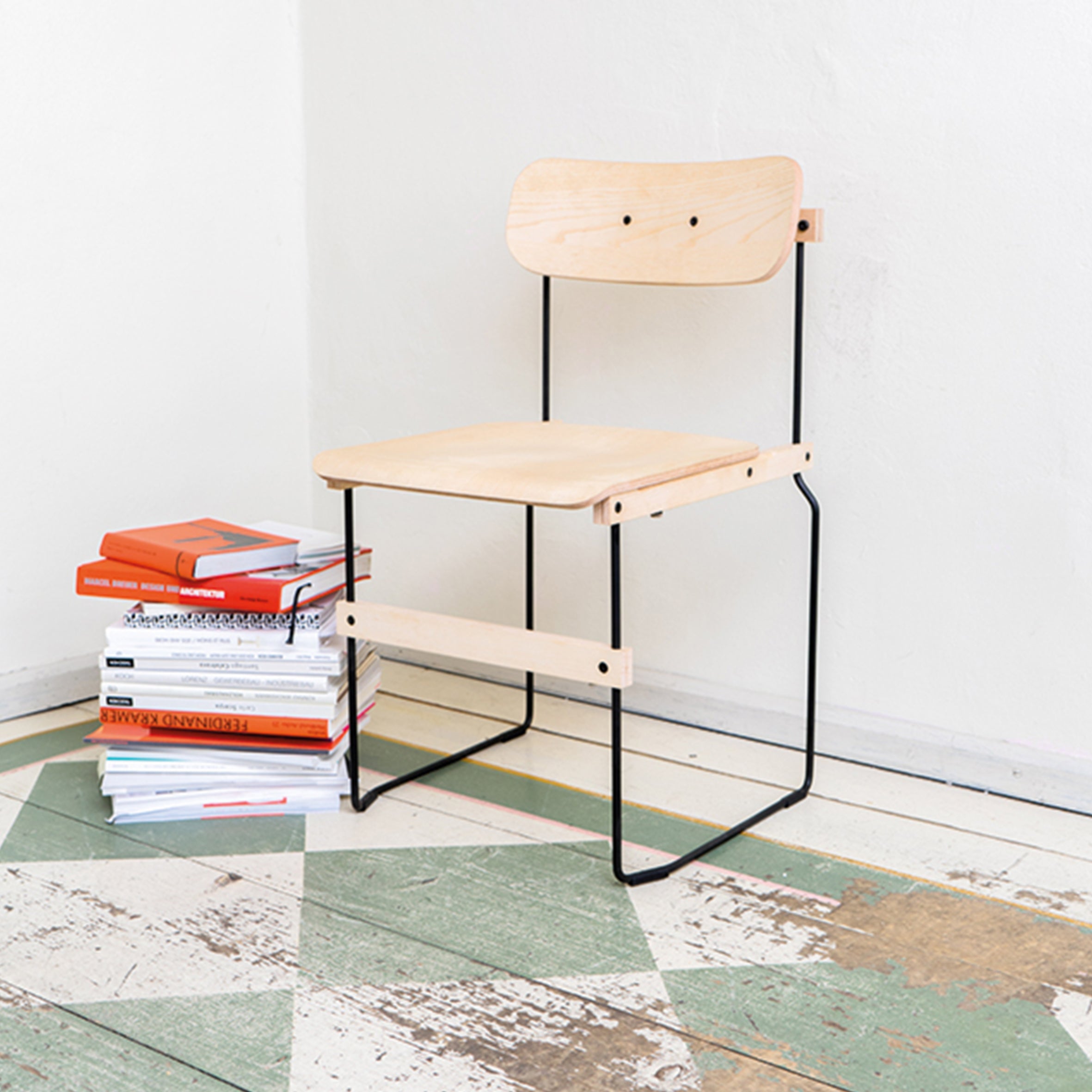 The Bruto chair by Moormann, with minimalist molded plywood design and a black metal frame, sits on a geometric-patterned floor next to a colorful stack of books. The rooms white walls enhance its simple, modern aesthetic.