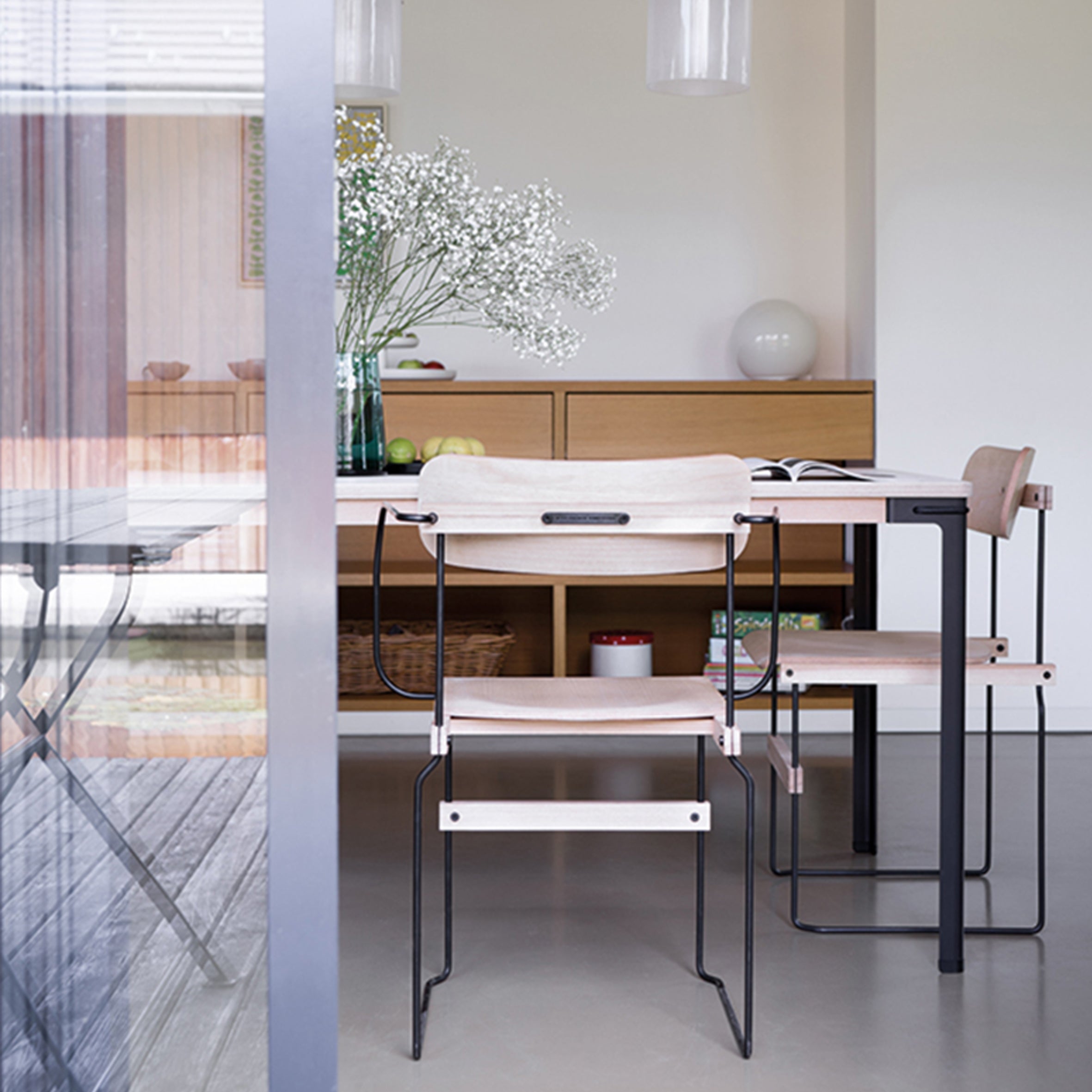 A modern dining area features a wooden table and chairs with black steel frames. A vase of white flowers accents the tabletop. In the background, a cabinet enhances the scene while a round lamp illuminates molded wood surfaces, with a glass door reflecting this flexible design. The Bruto Armchair by Moormann adds to the elegance.