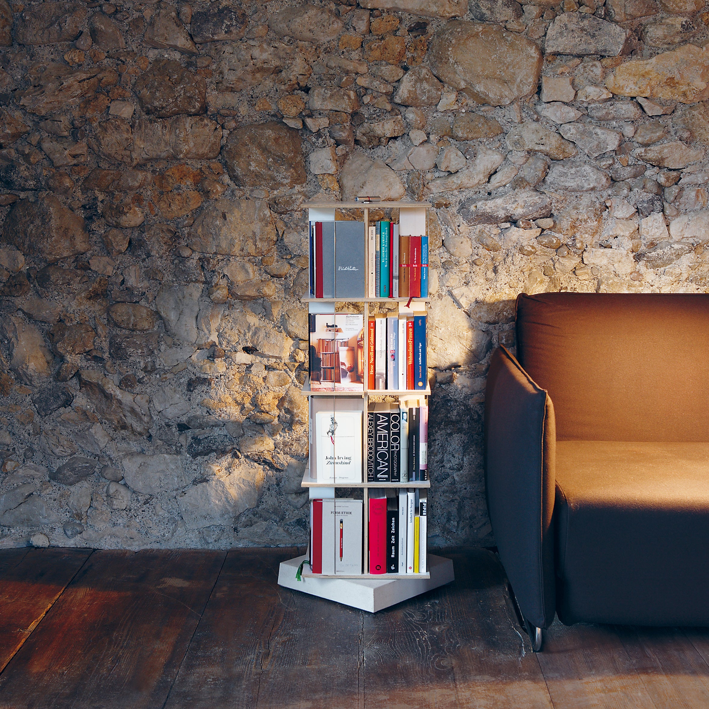 The Buchstabler by Moormann, a pivotable shelf holding various books, stands against a rustic stone wall. Its beside a brown sofa on a wooden floor, forming a cozy reading nook. This space-saving storage is bathed in soft lighting, casting a warm glow over the scene.