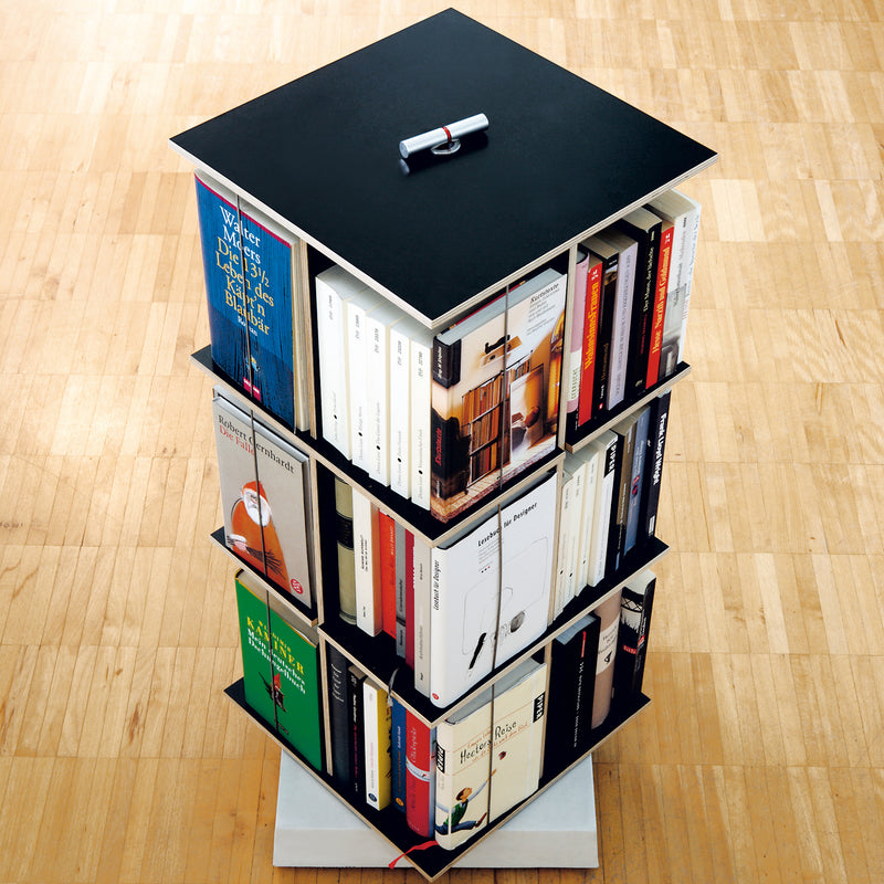 A space-saving, four-tiered Buchstabler bookshelf from Moormann, tightly packed with books of various sizes and colors, rotates on a light wooden floor. Its top is black with a handle.