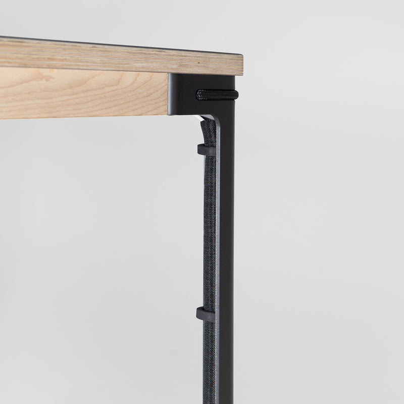 A close-up of the Moormann Cable Guidance table showcases a light wood tabletop with black metal legs and a black fabric wrap for cable management. Its minimalistic design features sleek magnetic brackets for an organized look against a white backdrop.