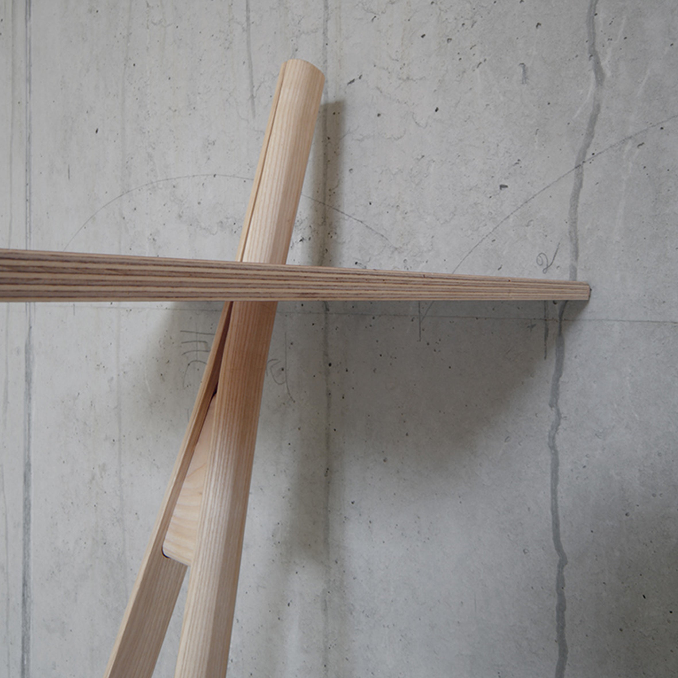 Against a light gray concrete wall, Moormanns Der Kleine Lehner—a minimalist wooden writing stand with angled beams—showcases clean lines and natural wood grain. This combination offers a space-saving, modern aesthetic that embodies elegance against the raw concrete backdrop.