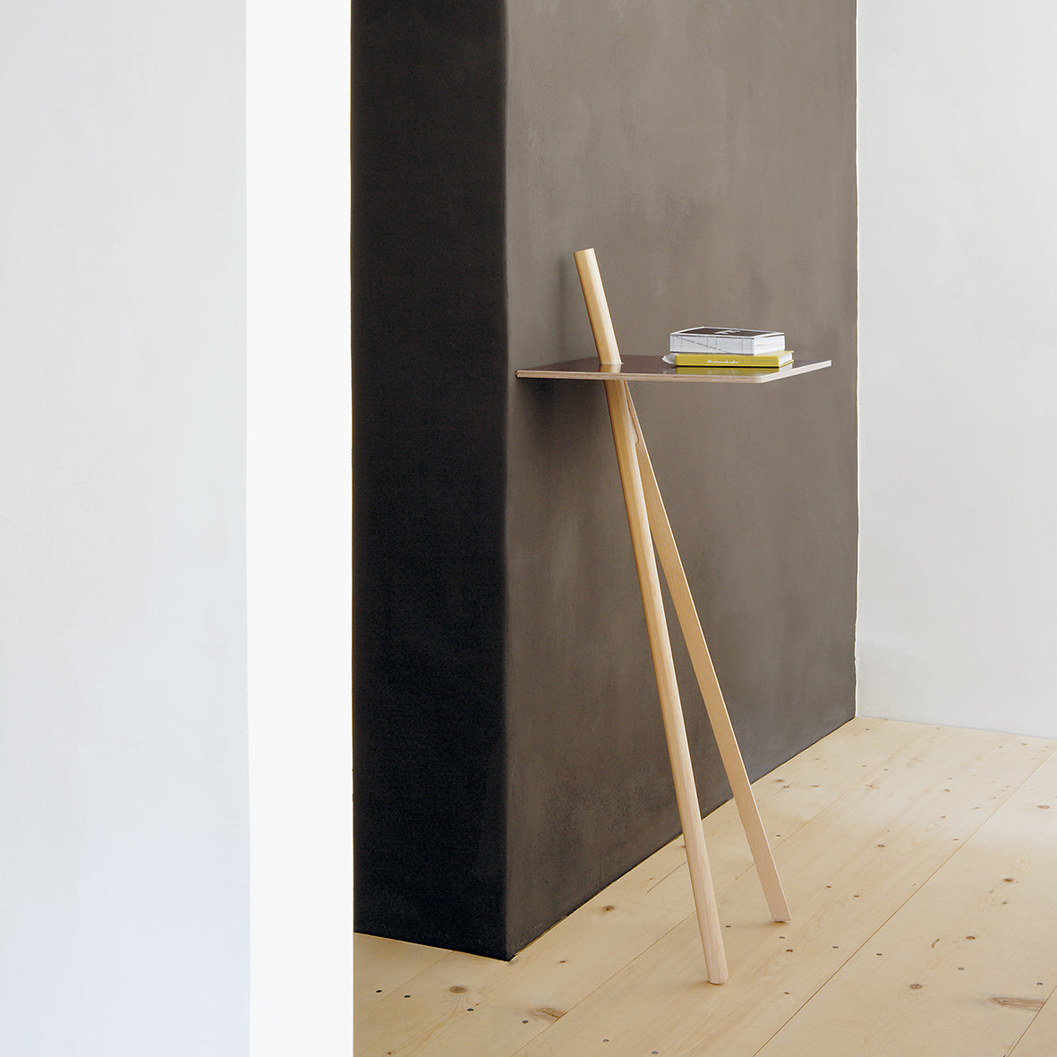 Check out the Der Kleine Lehner by Moormann: a minimalist leaning shelf with stylish wooden legs, perfect against a dark wall. It efficiently holds books while the light wood floor adds contrast, offering a sleek and modern look.