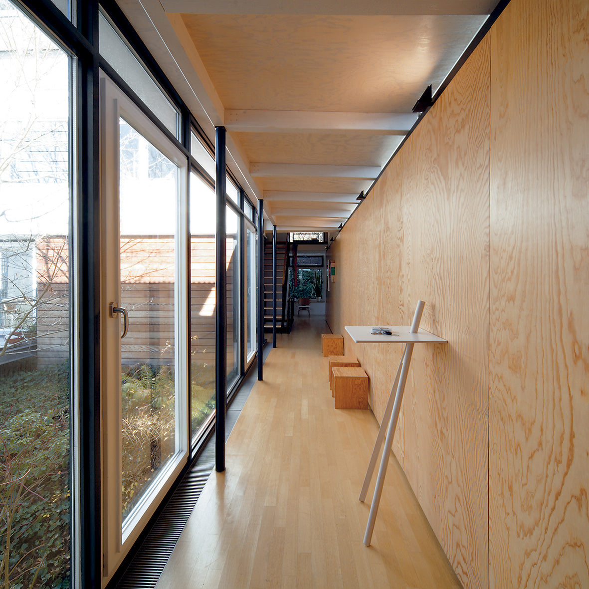A modern interior walkway with wooden floors and walls features two box-shaped wooden stools, a minimalist white table doubling as a space-saving writing stand for a tablet, and large glass doors on the left showcasing Moormanns Der Kleine Lehner garden, leading to a staircase in the background.