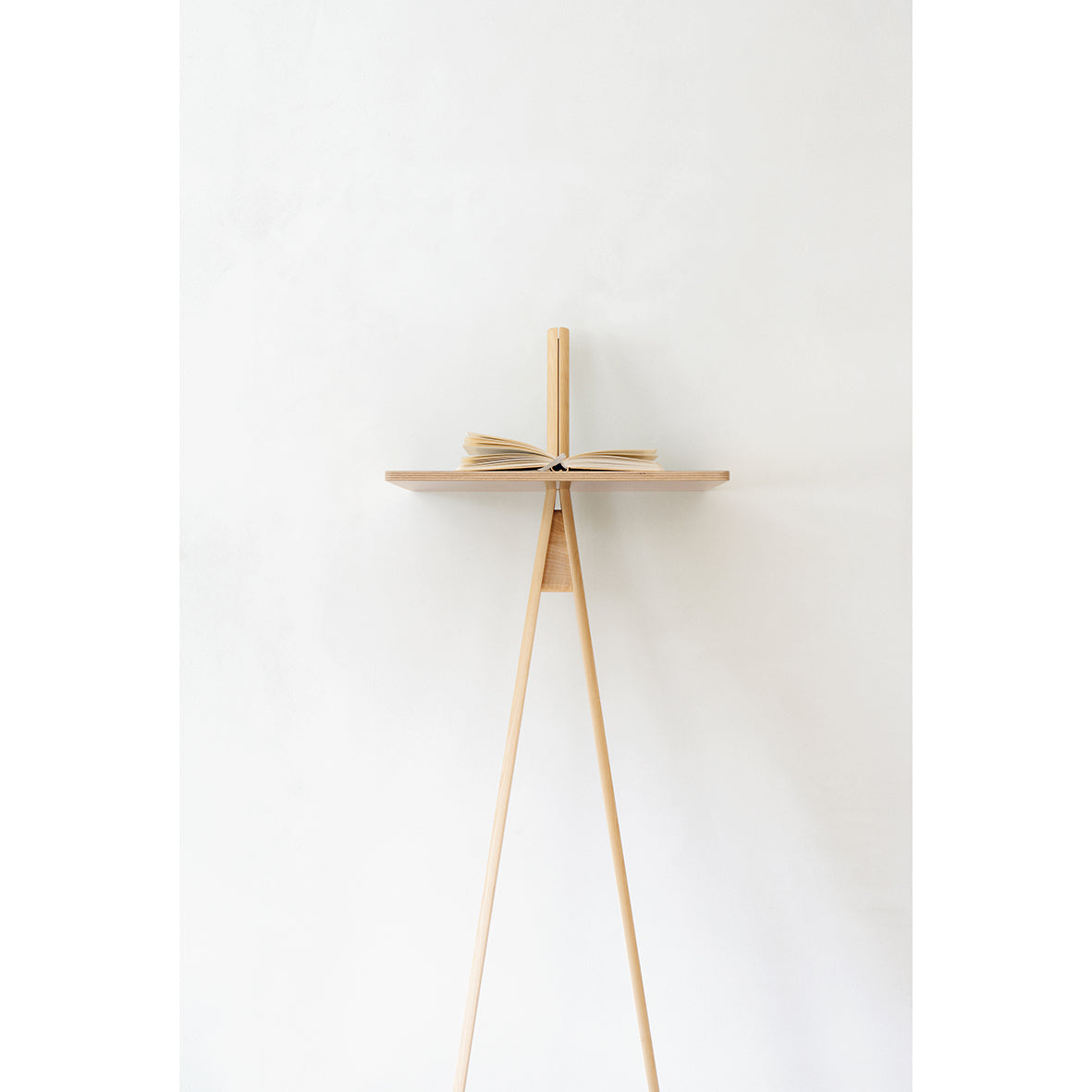 The Der Kleine Lehner by Moormann is a minimalist wooden book stand with three legs, supporting open books on a plain white background. Its simple, elegant design highlights natural wood tones and saves space efficiently.