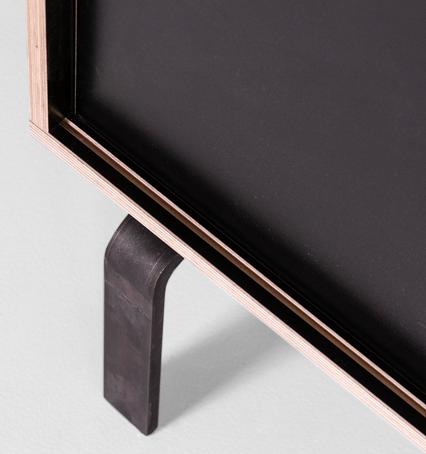 Close-up of the Egal Lowboard by Moormann, featuring a minimalist black tabletop and sleek leg resembling a refined sideboard. The layered wood frame showcases its natural grain, while the simple modern design contrasts dark and light materials effectively.