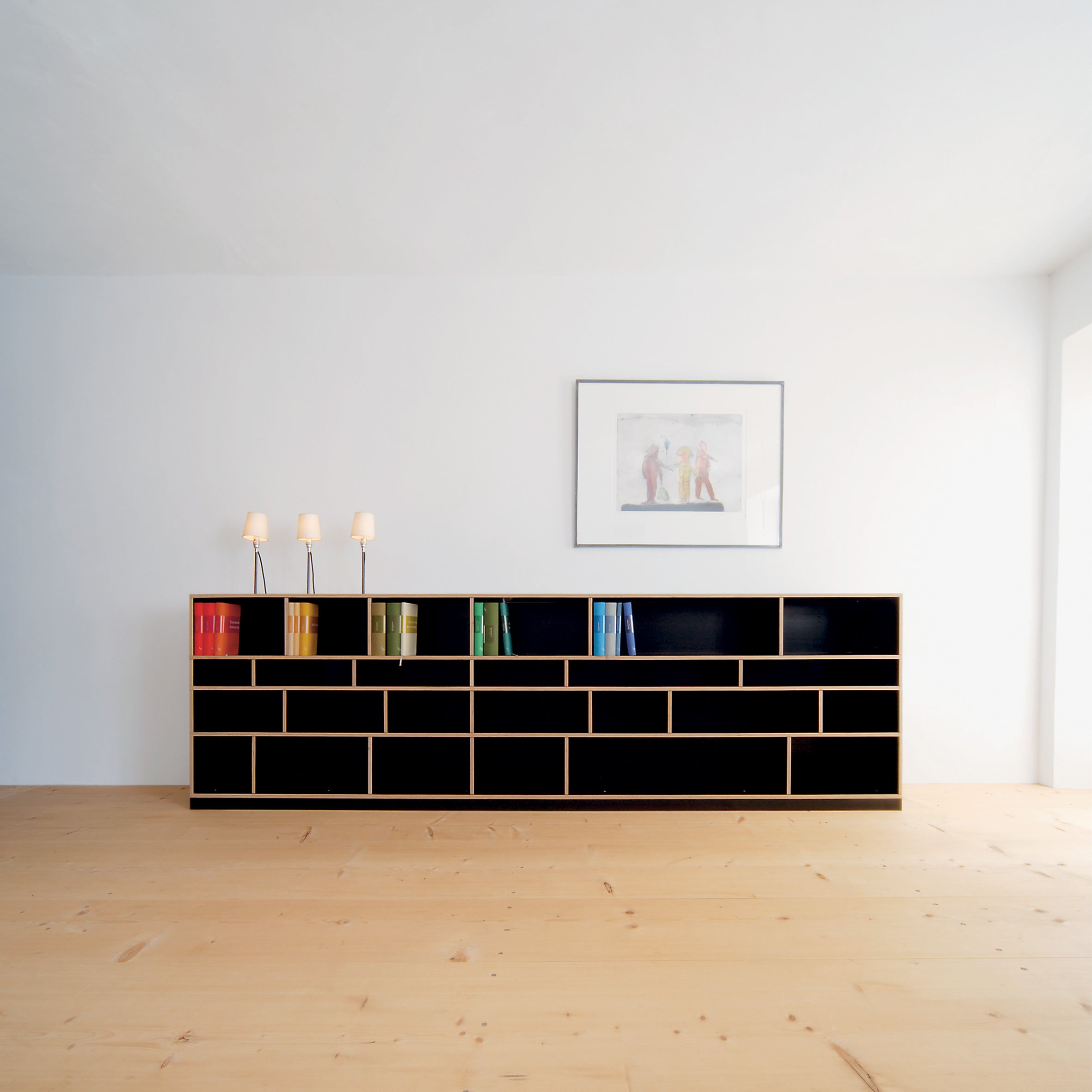 A minimalistic room showcases a black, brick-patterned Moormann bookshelf against a white wall. Rainbow-hued books add color, while the Rosi Lamp illuminates the space. Framed artwork hangs above, and the plain wooden floor ties together the elegant decor.