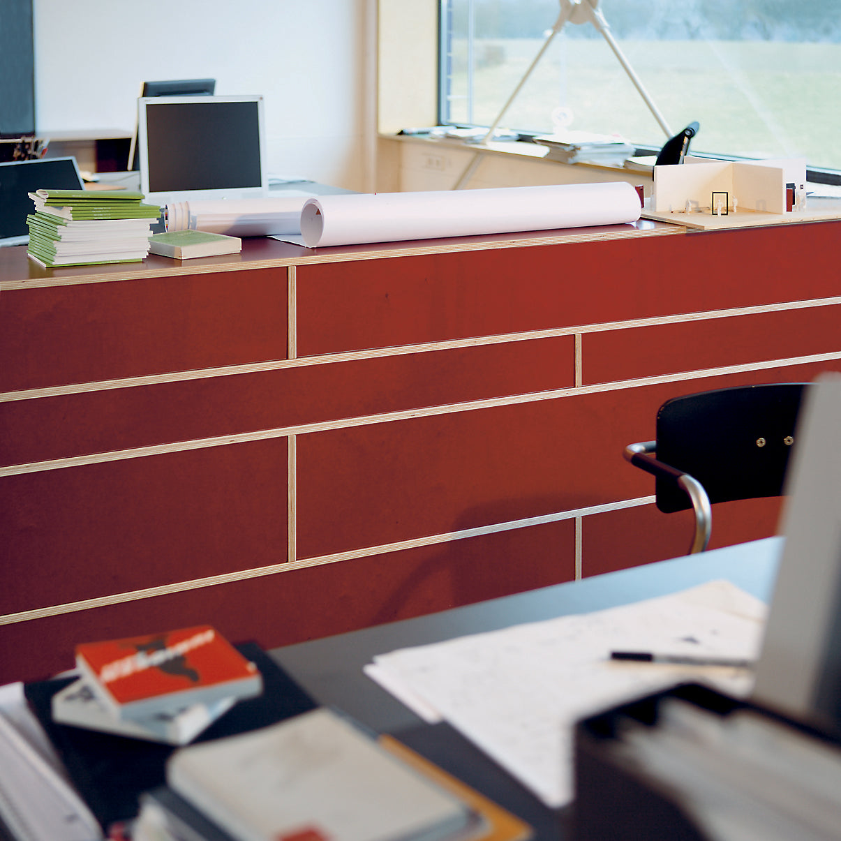 The modern office features a red desk with stacked papers, books, and a computer monitor. Nearby is a chair alongside plans or blueprints. Sunlight from a large window highlights the green view outside, while Moormanns Egal shelf system provides customizable storage for organization.