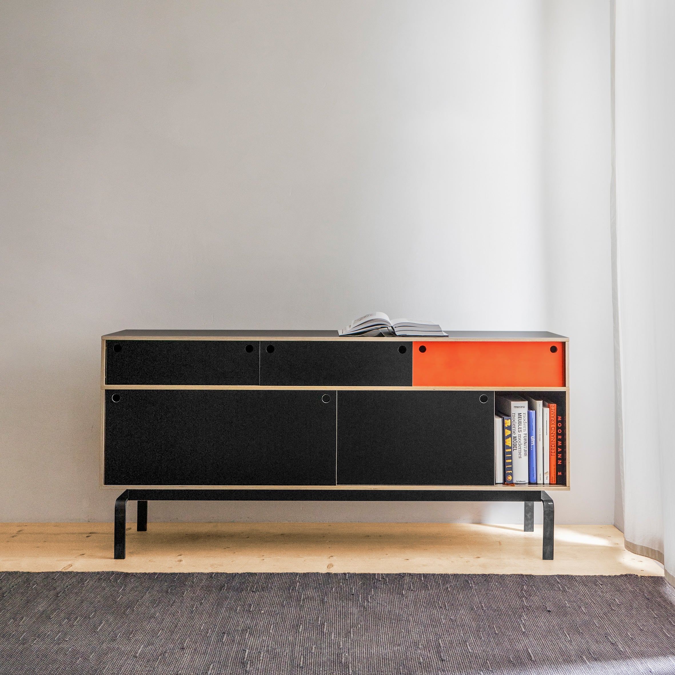 The Moormann Egal Sideboard, made from birch plywood with black and orange accents, is set against a light gray wall. It features sliding doors and open compartments that hold books and a cup, elegantly placed on a dark rug over a light wooden floor.