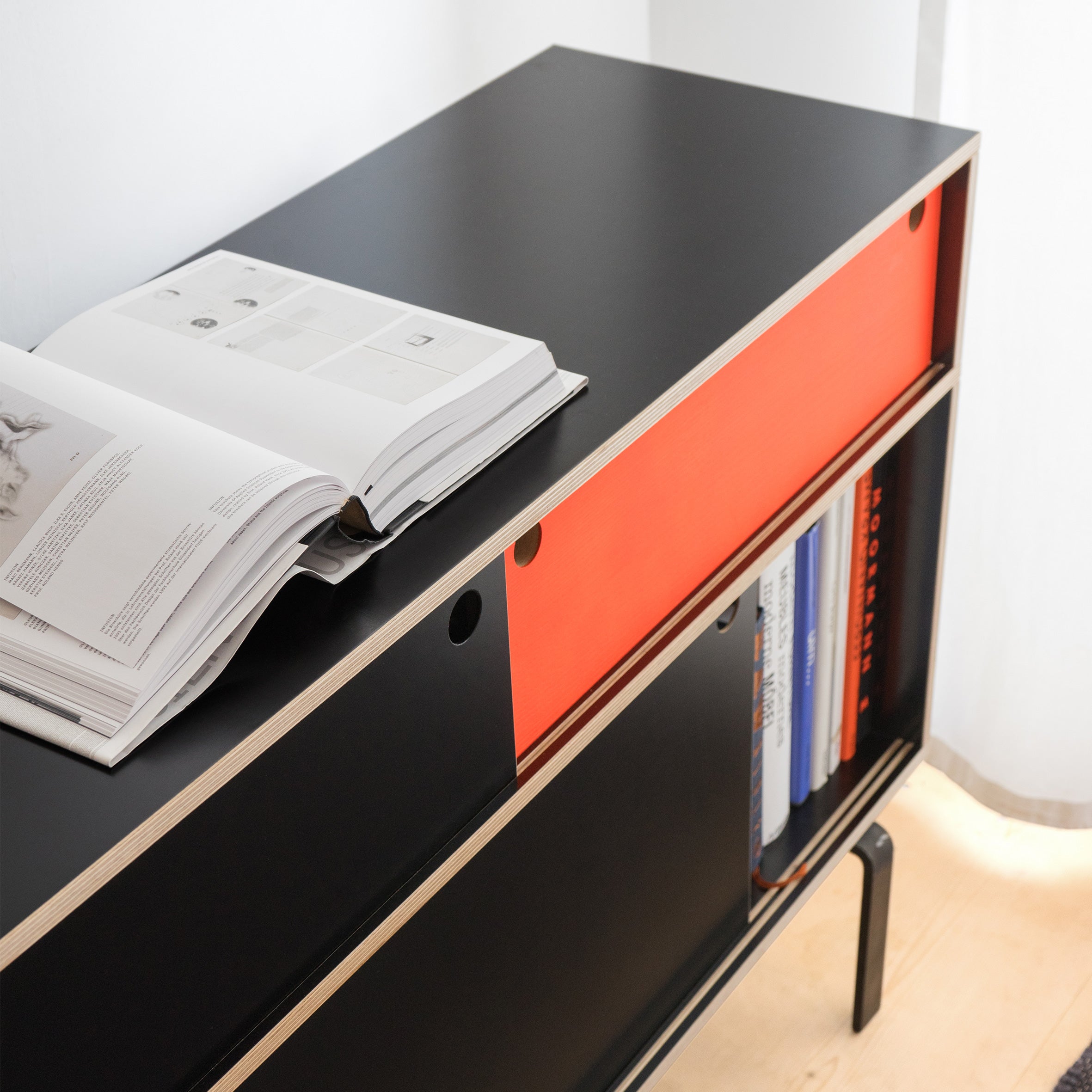 In a well-lit room, the Moormann Egal Sideboard, crafted from sleek birch plywood with modern black and red sliding doors, is topped by an open book. Neatly arranged books on the lower shelf add style and functionality.