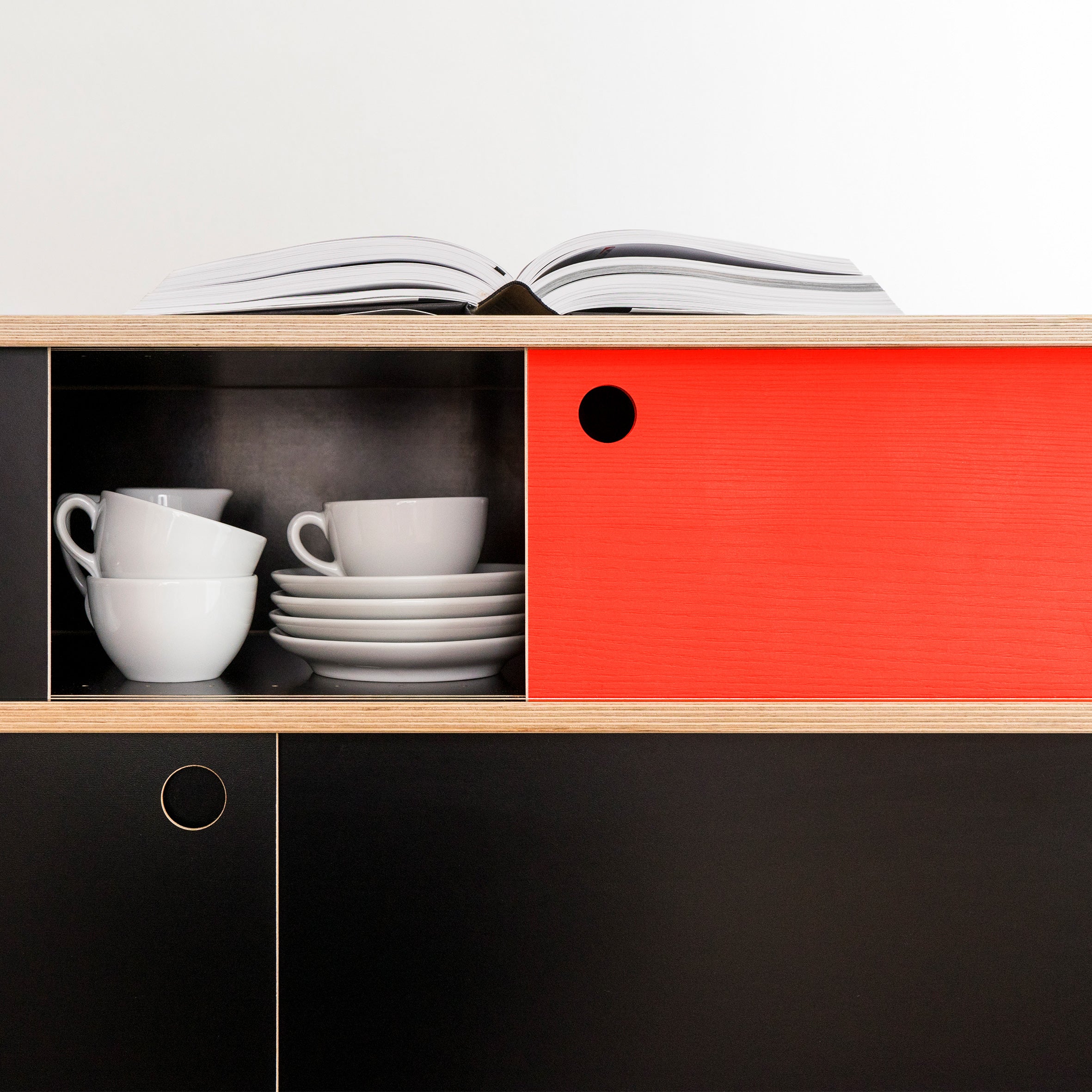 A Moormann Egal Sideboard made from birch plywood showcases a minimalist design with a red sliding door, partially open to display white cups and saucers. An open book on top complements the sleek, modern look of the neutral space.