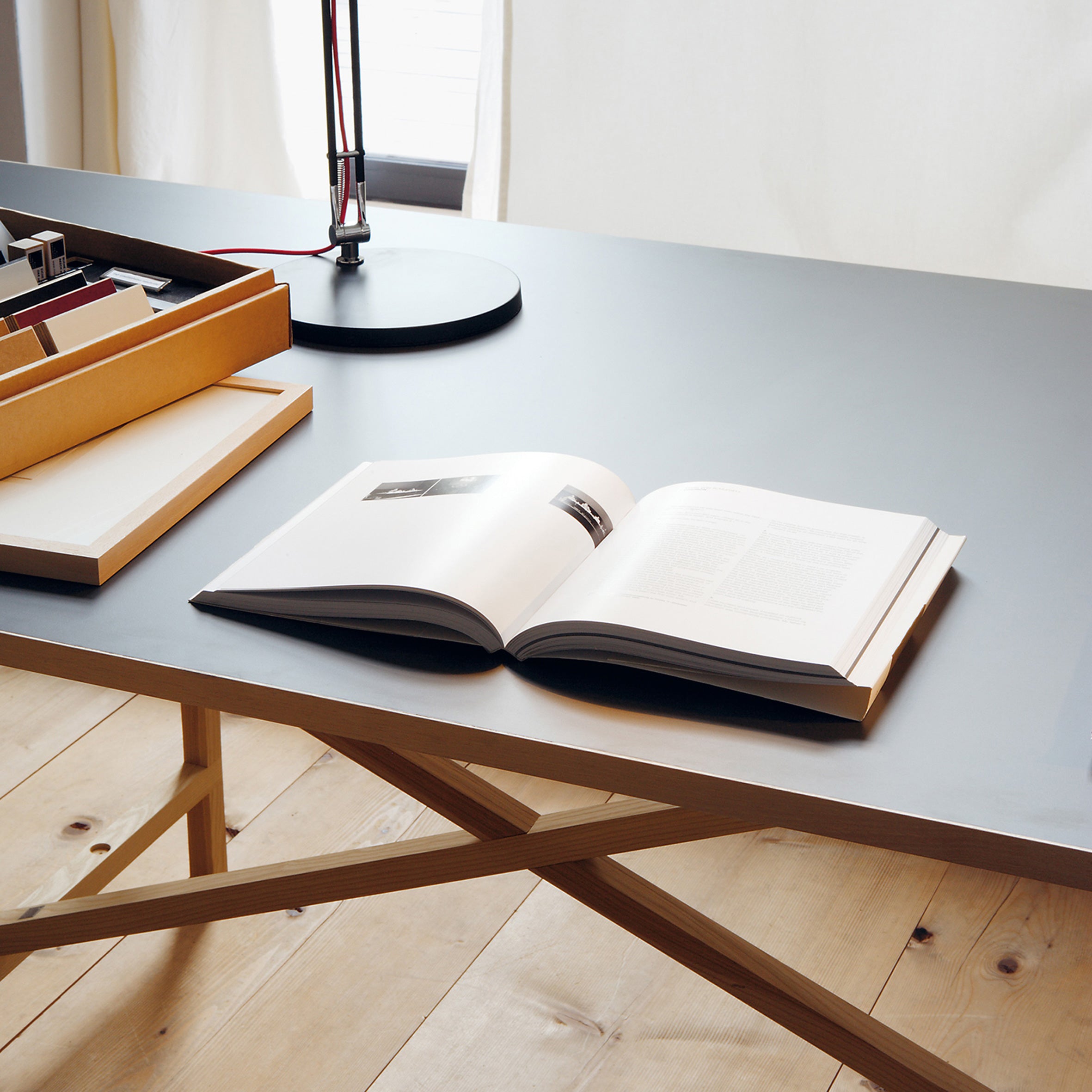 The Egon desk by Moormann boasts a modern, minimalist design with a light wooden frame and grey top free of metal. An open book, closed cardboard box, and sleek black lamp create a calm, organized atmosphere in this well-lit setting.