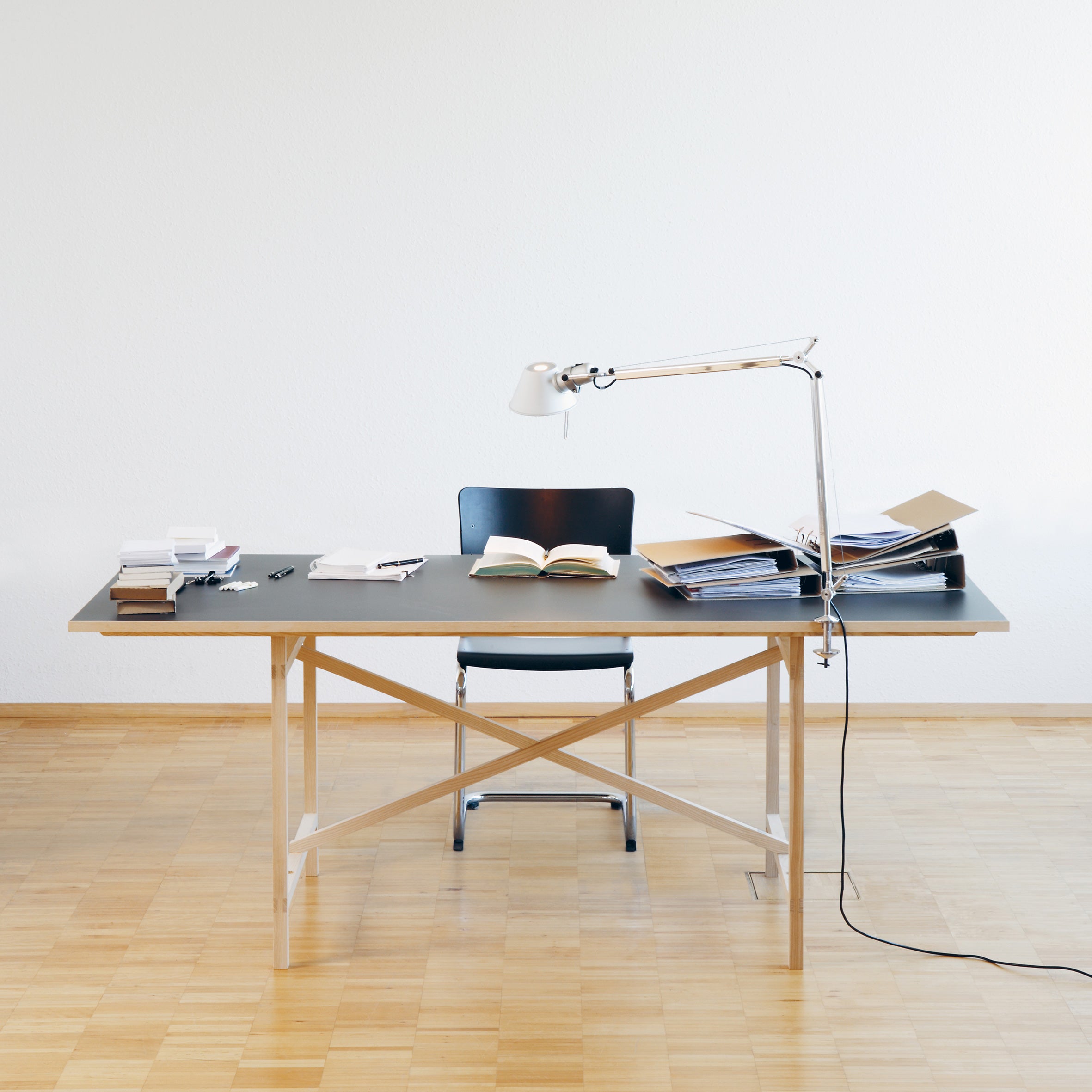 A minimalist office setup with Moormanns Egon includes a wooden desk, black chair, and sleek lamp. Open books, files, and pens are scattered on the table. This arrangement against a plain white wall and wooden flooring reflects Franz Volhards metal-free design style for an organized workspace.