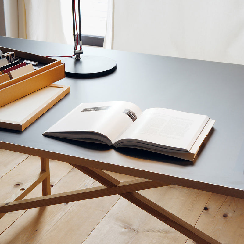 The Egon desk by Moormann boasts a modern, minimalist design with a light wooden frame and grey top free of metal. An open book, closed cardboard box, and sleek black lamp create a calm, organized atmosphere in this well-lit setting.