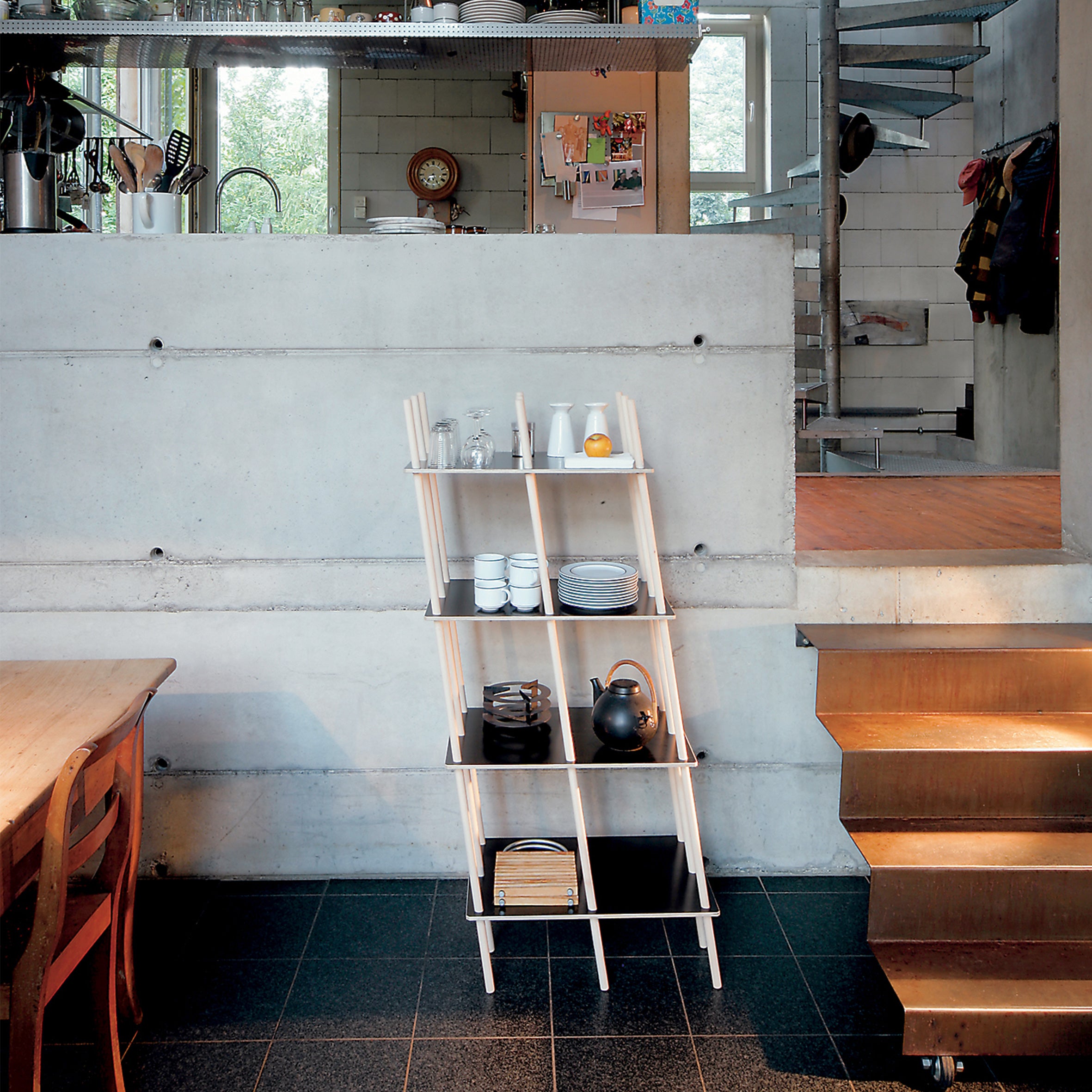 In a modern kitchen, a Moormann Es minimalist leaning shelf adorns a concrete wall, showcasing white dishes, teapots, and a lemon. Wooden steps ascend to an elevated area overlooking kitchen utensils and a window. A wooden table is partially visible.