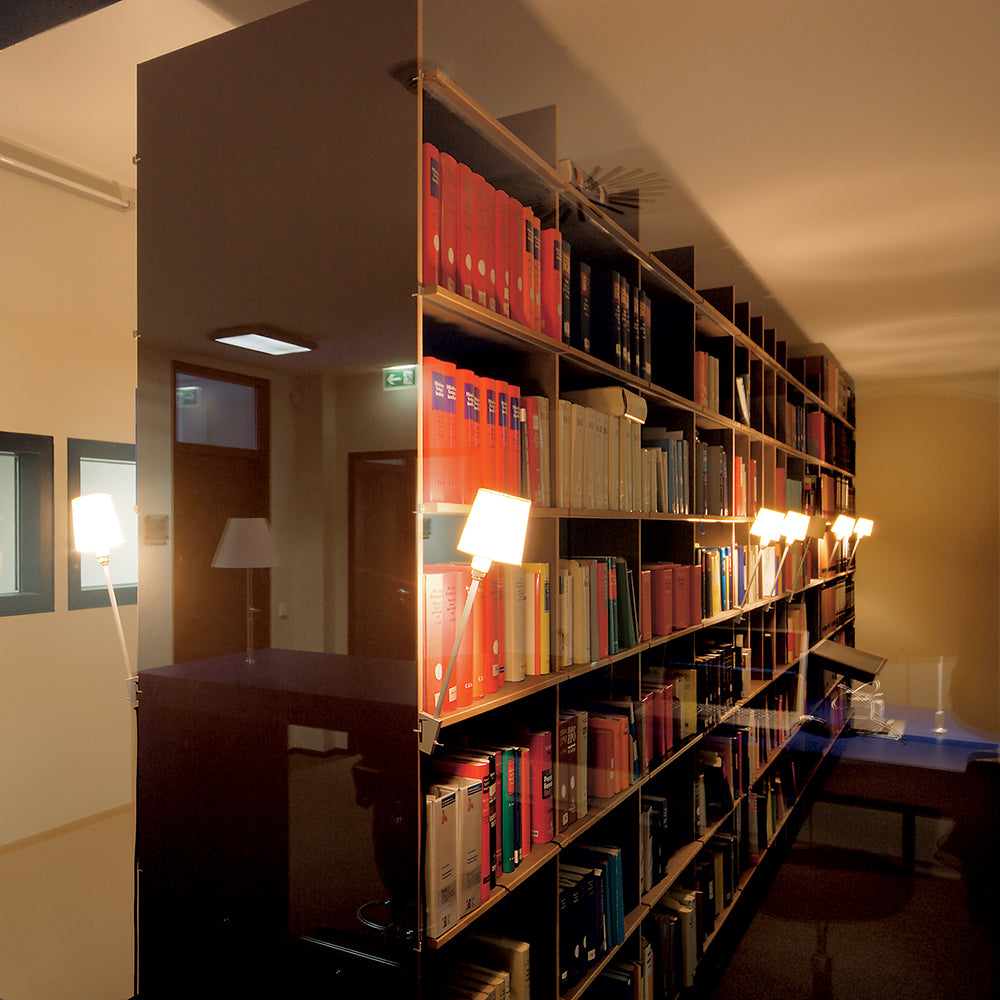 The contemporary library features the Moormann FNP Archive freestanding shelf, surrounded by other colorful book-filled shelves. Angular wall lights provide soft illumination, while a reflective glass wall on the left enhances the warm and inviting atmosphere.