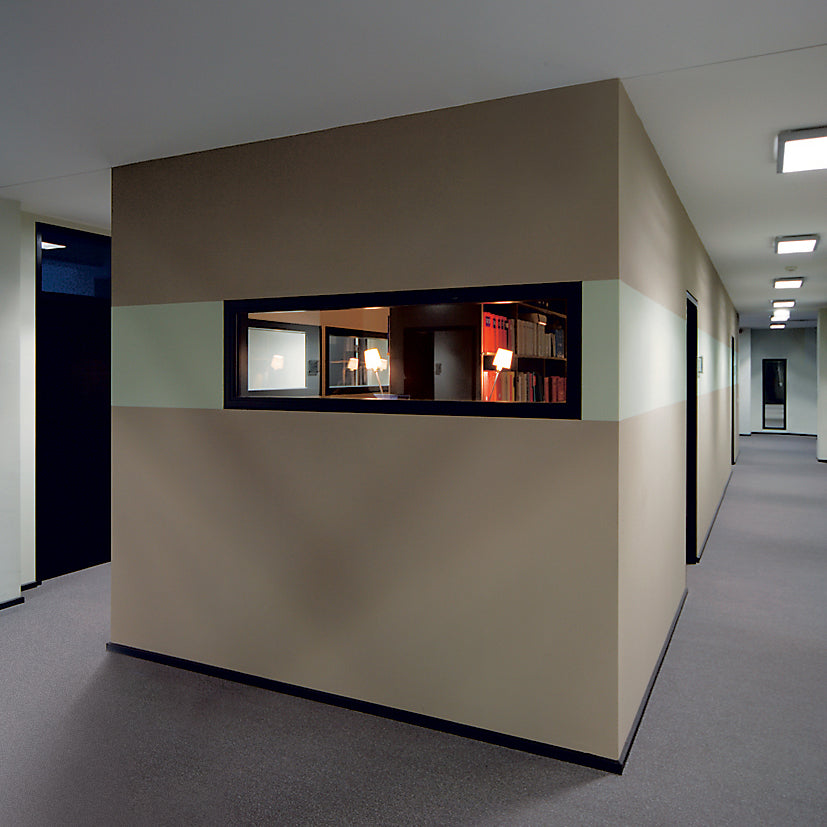 The minimalist office hallway features beige walls and a rectangular window offering a view of the modern workspace. Moormanns FNP Archive accompanies the lamp and freestanding shelf, with bookshelves, carpeted flooring, and ceiling lights enhancing the tidy, professional setting.