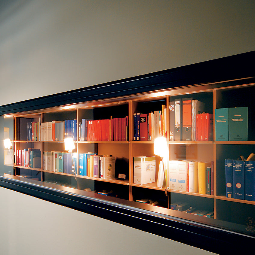 The Moormann FNP Archive is a long, wall-mounted shelf filled with colorful books. Two warm lights illuminate it, creating a cozy atmosphere. With a black border, this storage unit adds modern flair to any room.