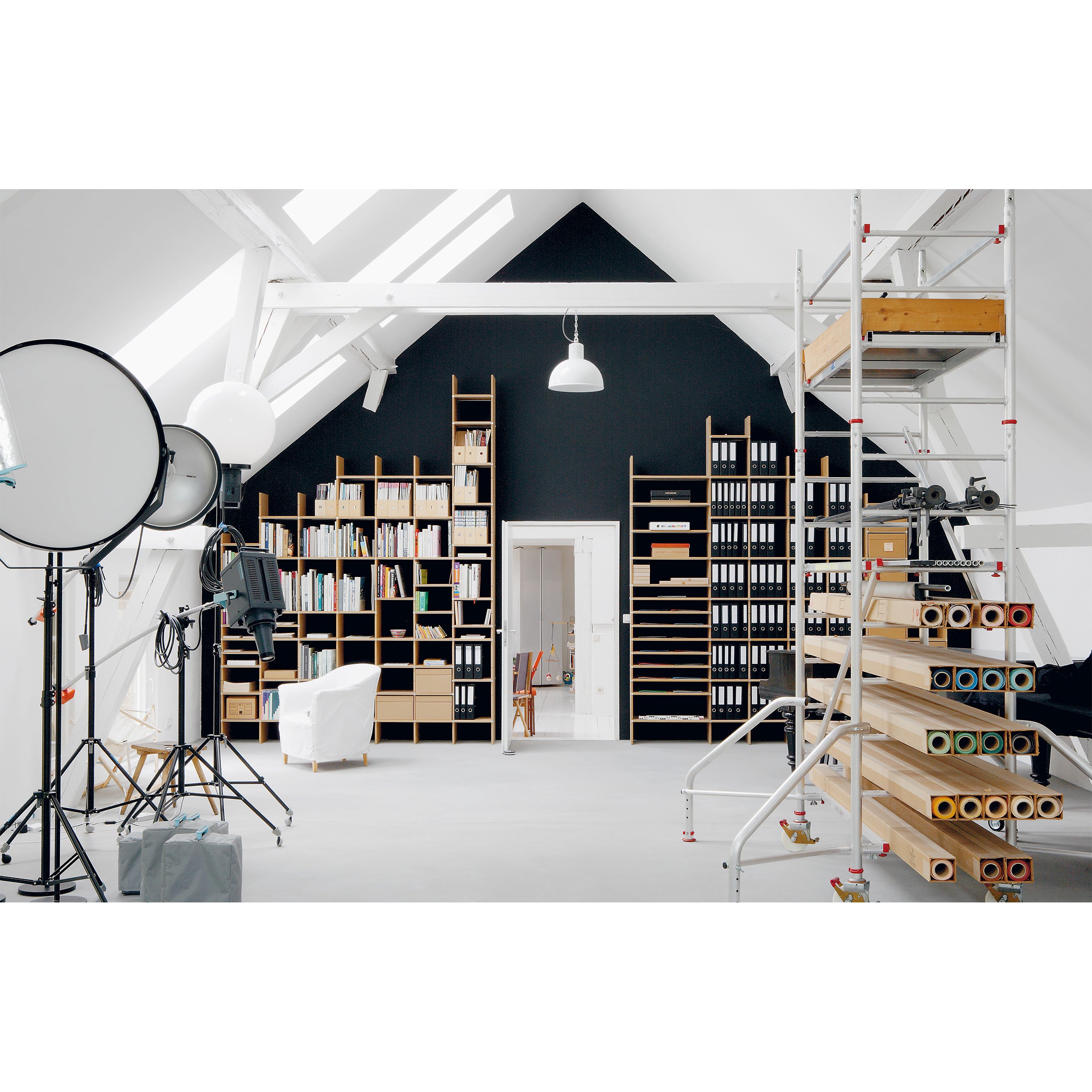 A spacious attic studio with adaptable design features white walls, ceiling beams, and FNP by Moormann shelves filled with books and folders. Large paper rolls and lighting equipment surround a cozy white armchair.