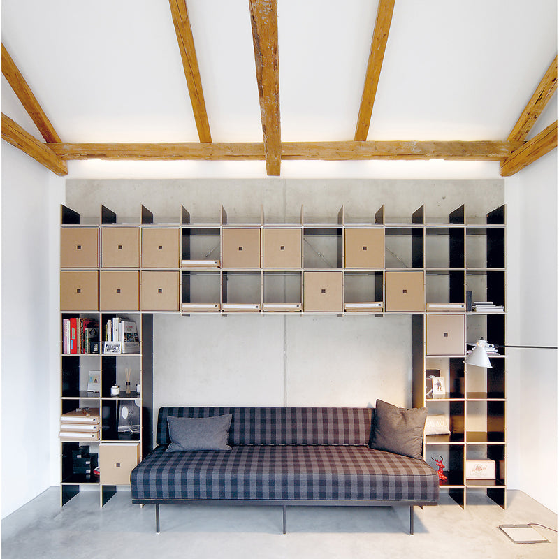 A modern living room features a checkered sofa with two cushions, accompanied by Moormanns adaptable FNP shelf on the wall. These units, both open and closed, showcase books and decor. Rustic wooden beams on the ceiling complete the look.