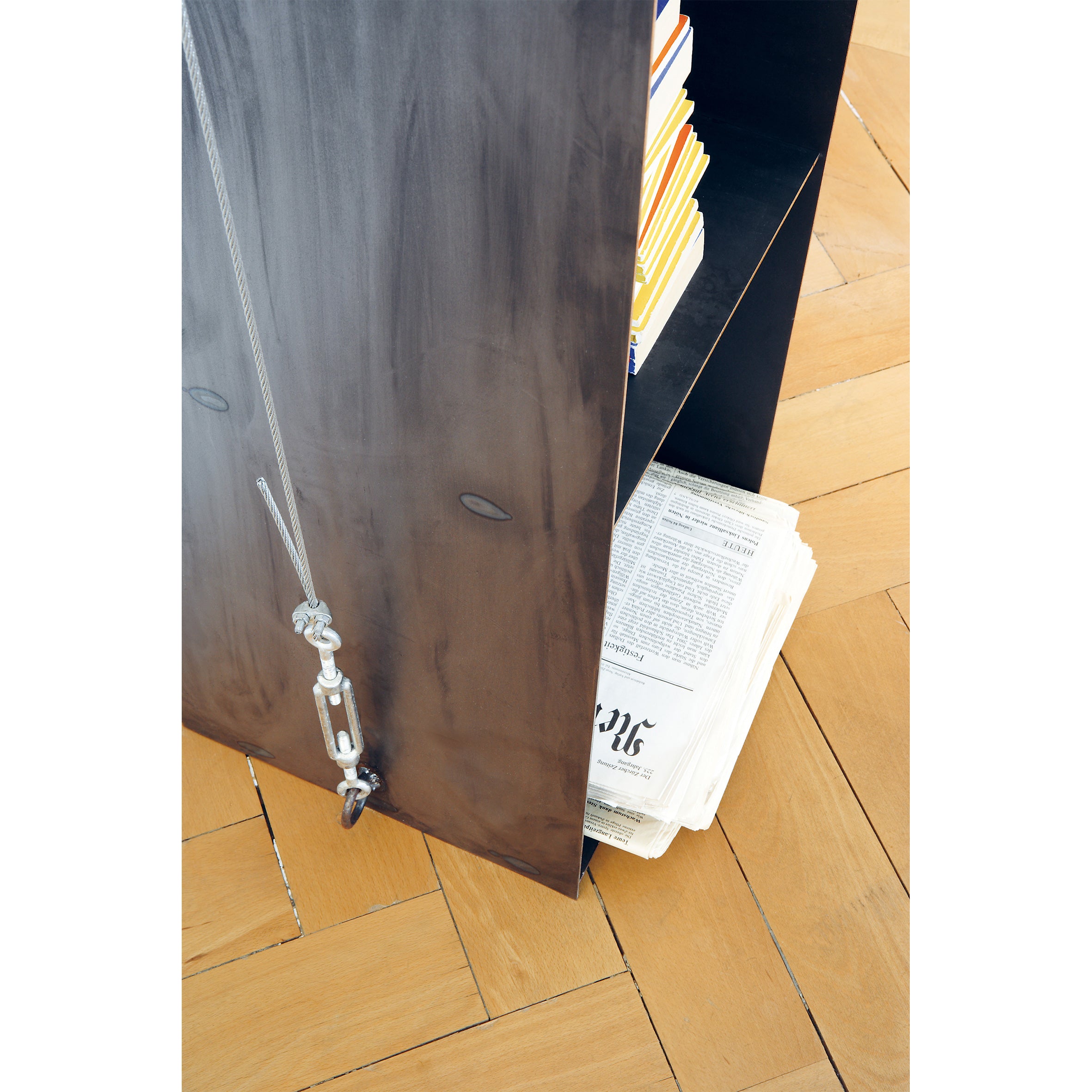 A sleek black bookshelf, similar to Moormanns Gespanntes Regal, features a side rope and hook. It elegantly displays books on the top shelf and newspapers on the bottom, all resting on a light wood parquet floor.