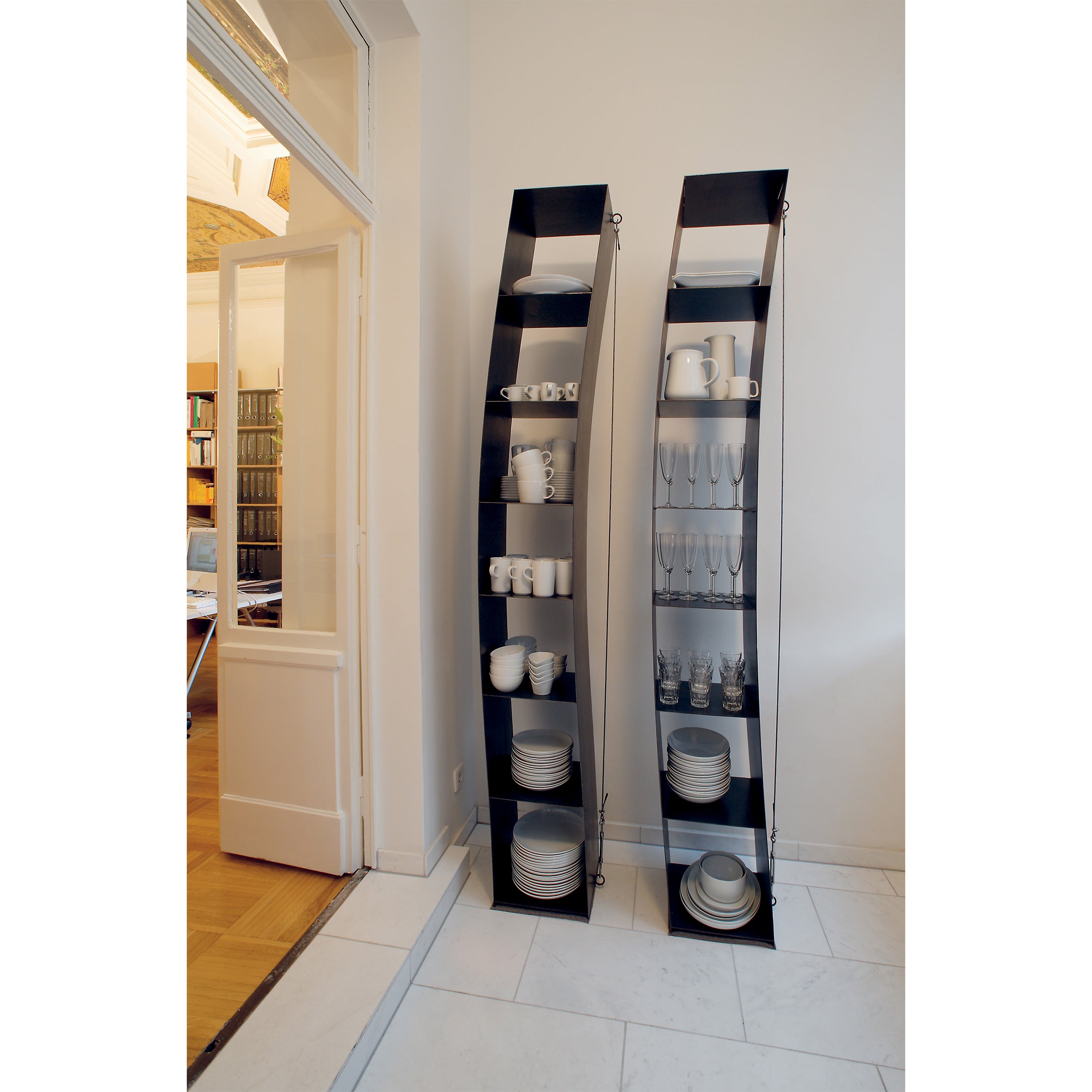 Two tall, slim black shelves, inspired by Moormanns elegant Gespanntes Regal, hold neatly arranged white dishes and glassware against a white wall in the bright room that leads into an area with more bookshelves and furniture.