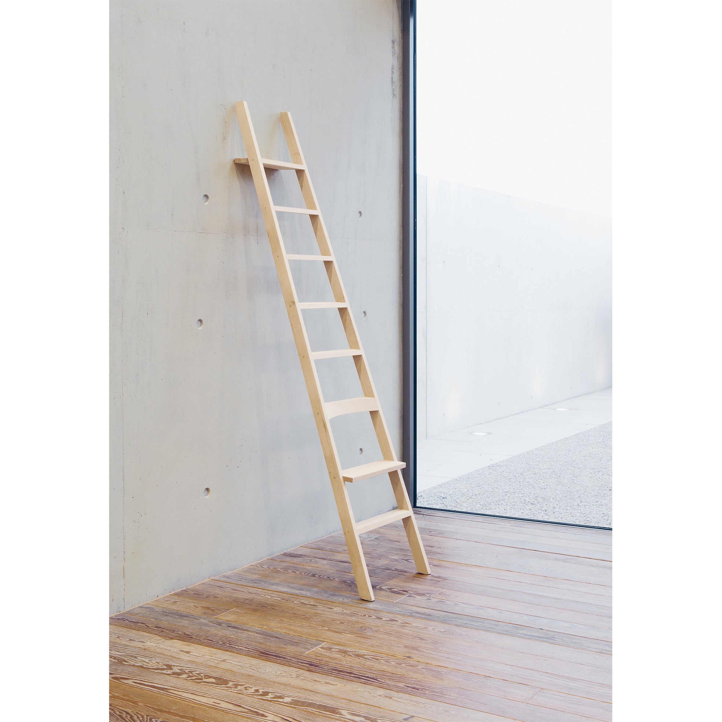 The Hochacht ladder by Moormann stands against a light concrete wall beside a large window. The room, with its wooden floor, reveals an outdoor gravel area awash in soft, diffused lighting.
