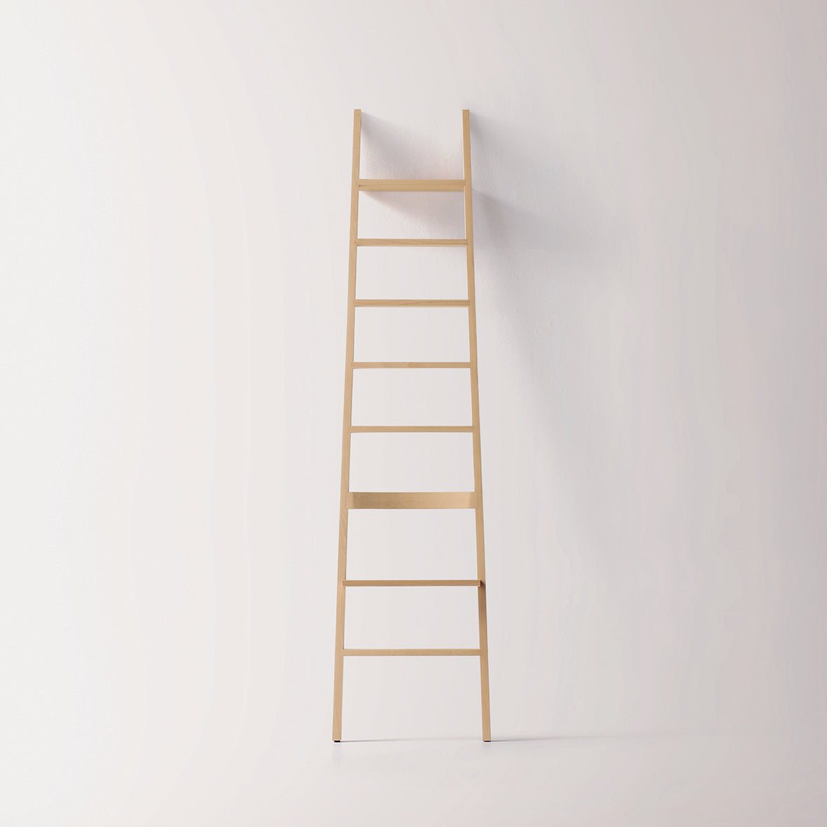 A Moormann Hochacht minimalist wooden ladder with a sleek, narrow design leans against a plain white wall. Its light beech untreated finish enhances the rooms airy, clean aesthetic.