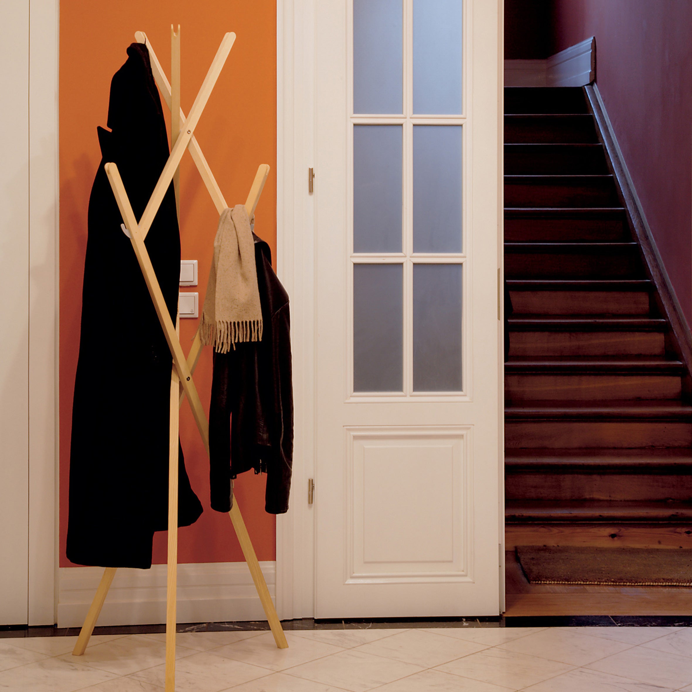 The Hut ab coat rack by Moormann stands against an orange wall with a black coat and beige scarf. A white glass-paneled door leads to a wooden staircase, creating a space-saving arrangement that complements the light-tiled floor beautifully.
