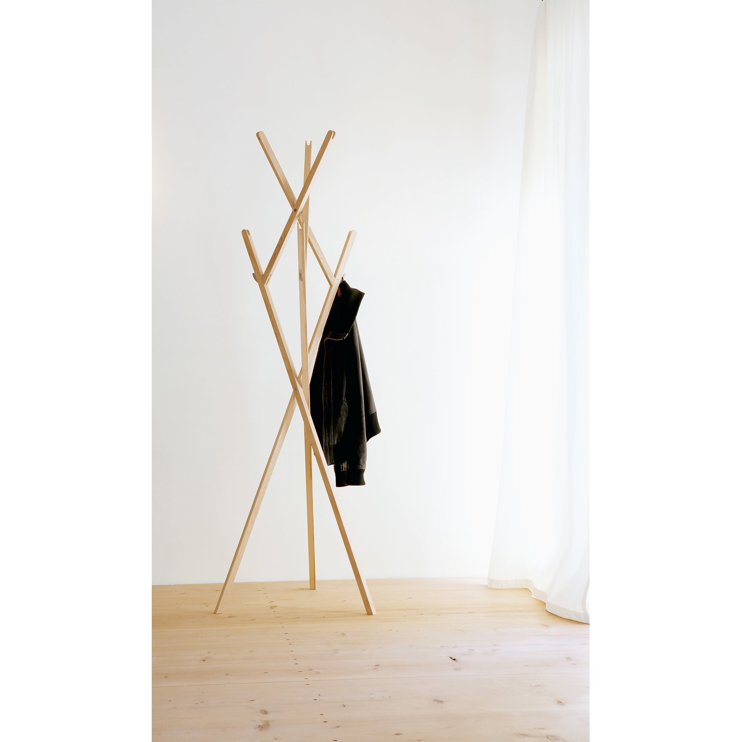 Moormanns minimalist Hut ab coat rack, featuring an asymmetrical design, holds a single black jacket on a light wooden floor. In the background, a sheer white curtain hangs against a plain white wall.
