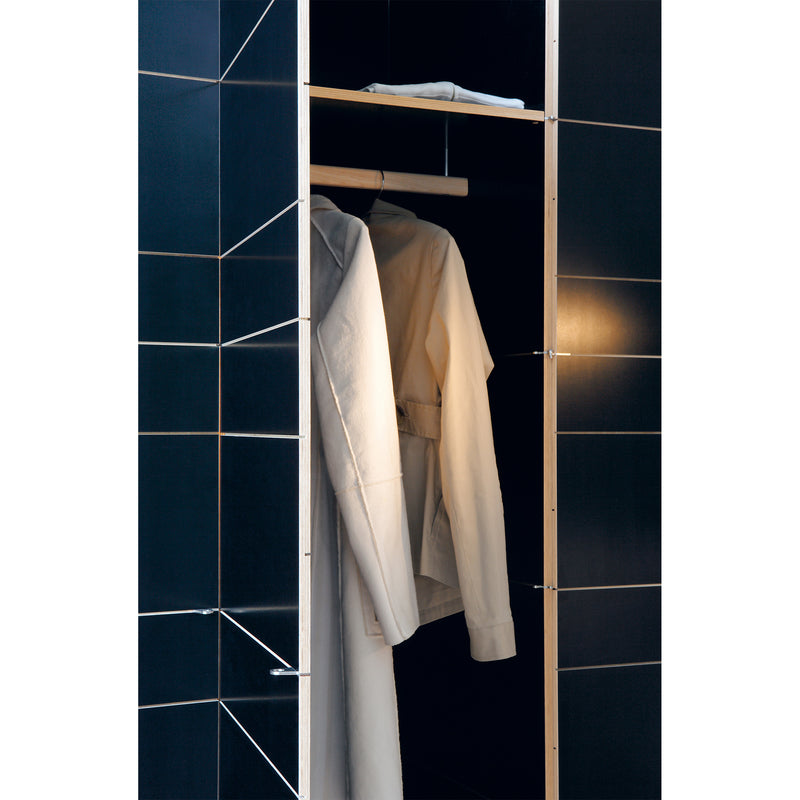 A partially open, black-tiled closet features a beige coat hanging inside on a wooden rod, highlighting Moormanns sleek K1 Storage modular system with the door ajar, offering a glimpse of the minimalistic interior.