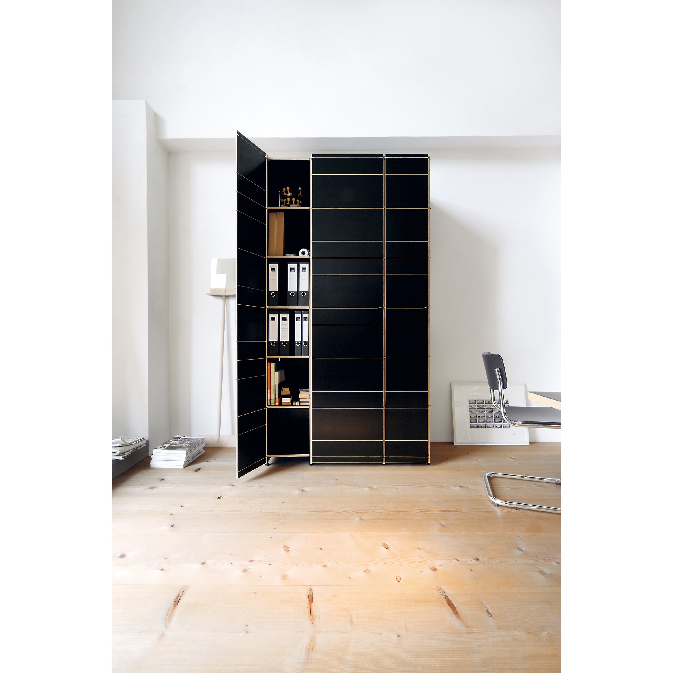 A modern office showcases a black K1 Storage cabinet by Moormann with one door open to reveal its modular system, neatly organizing binders and supplies. A sleek metal chair is placed by a small desk, all set on a light wooden floor.